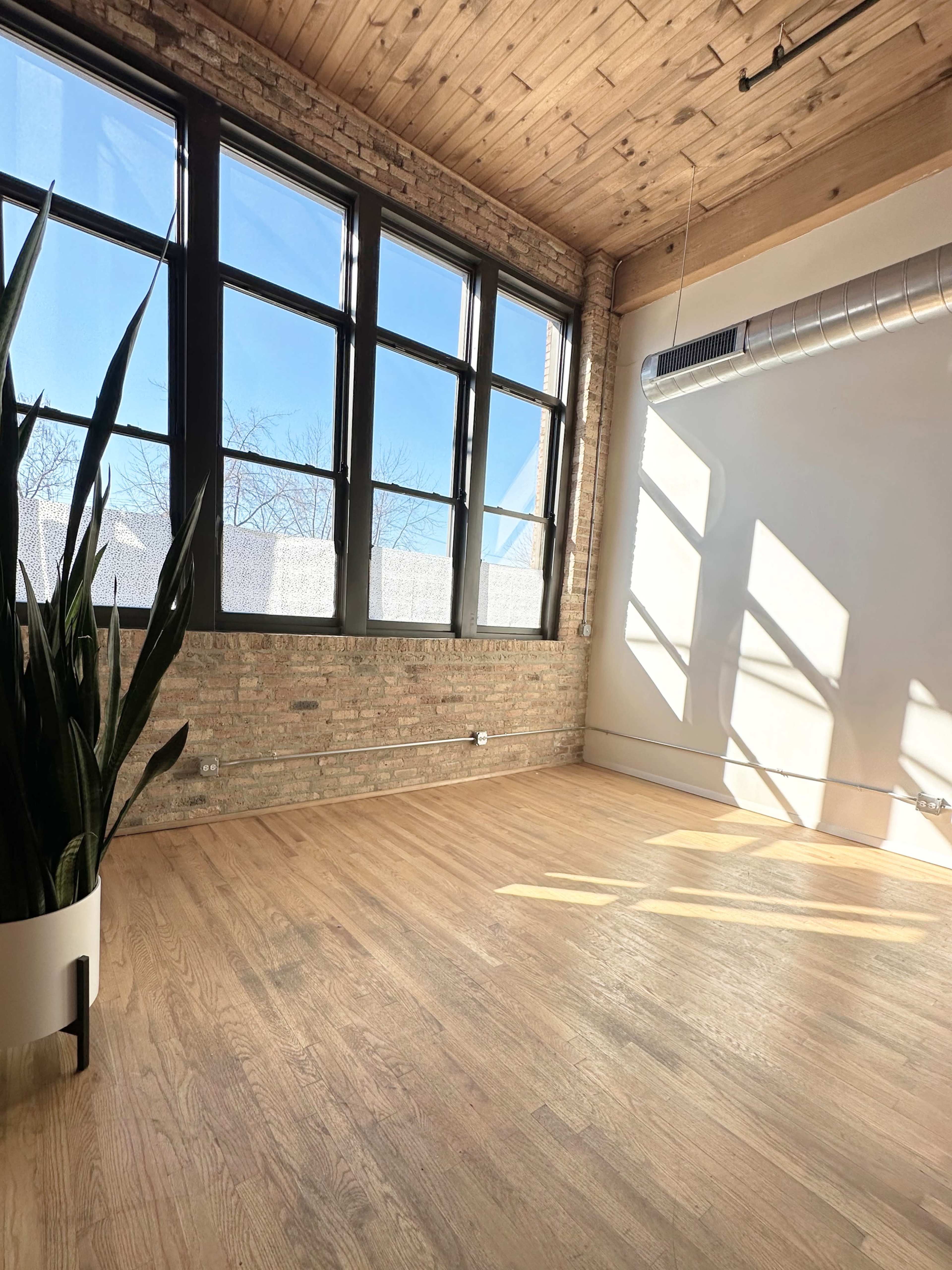 An empty room with large windows allowing natural light to fill the space, featuring exposed brick walls and hardwood floors.