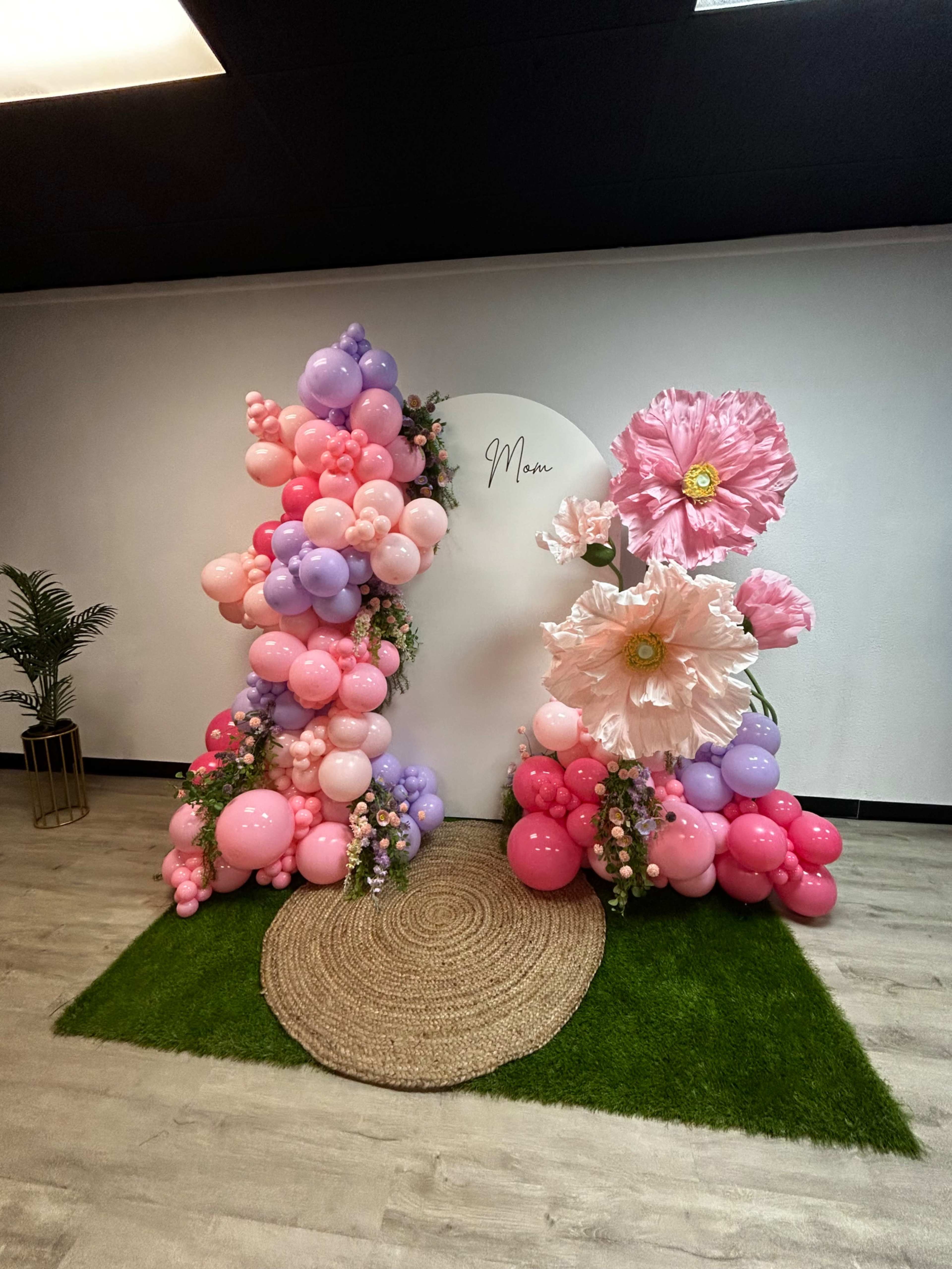 The image shows a festive backdrop decorated with pink and purple balloons and large artificial flowers, set against a white wall and a circular sign labeled "Mom," with a green grassy area and a round woven rug on the floor.