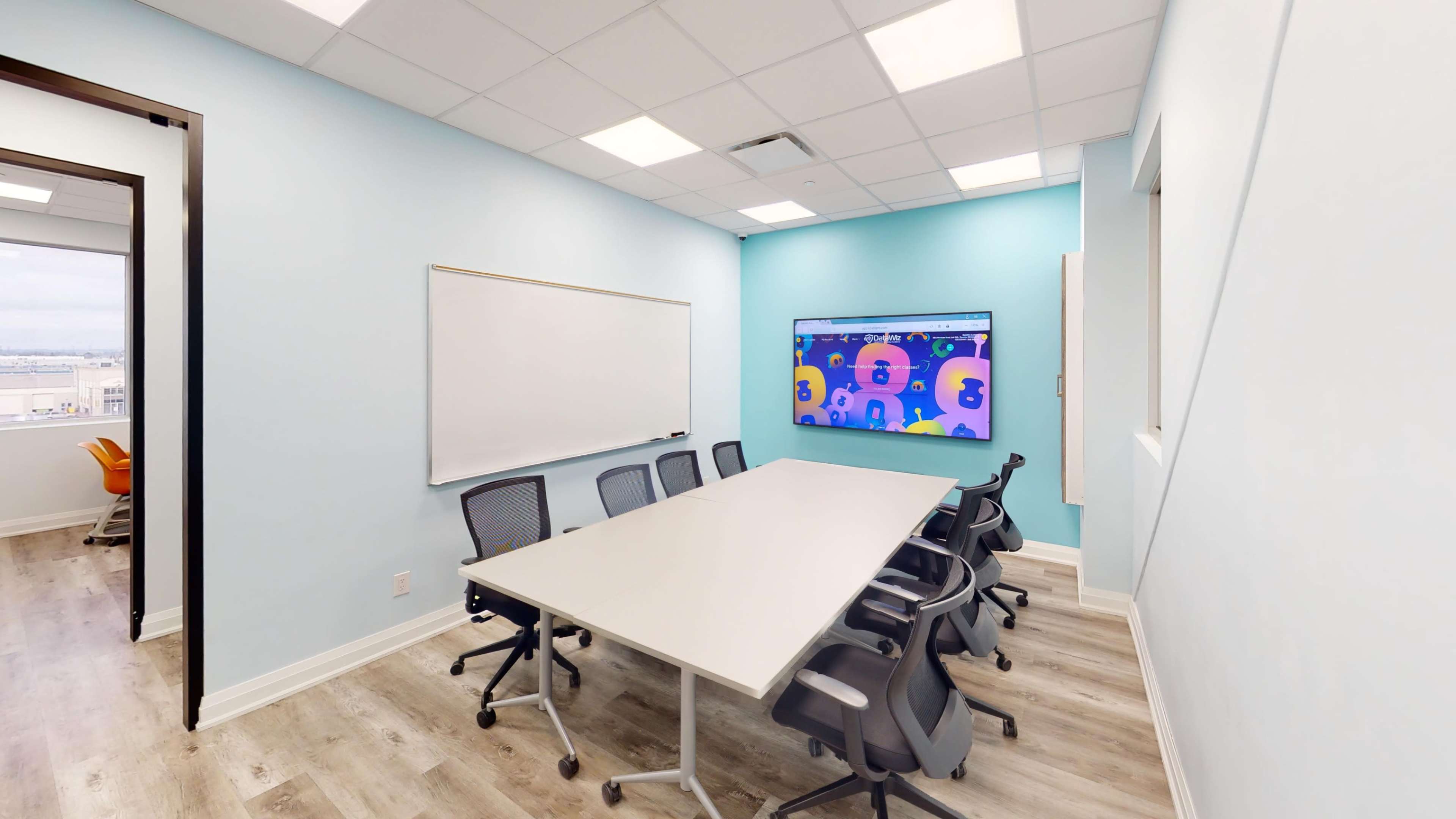 A meeting room features a long white table surrounded by ergonomic chairs, a whiteboard, and a large screen displaying colorful graphics on a light blue wall.