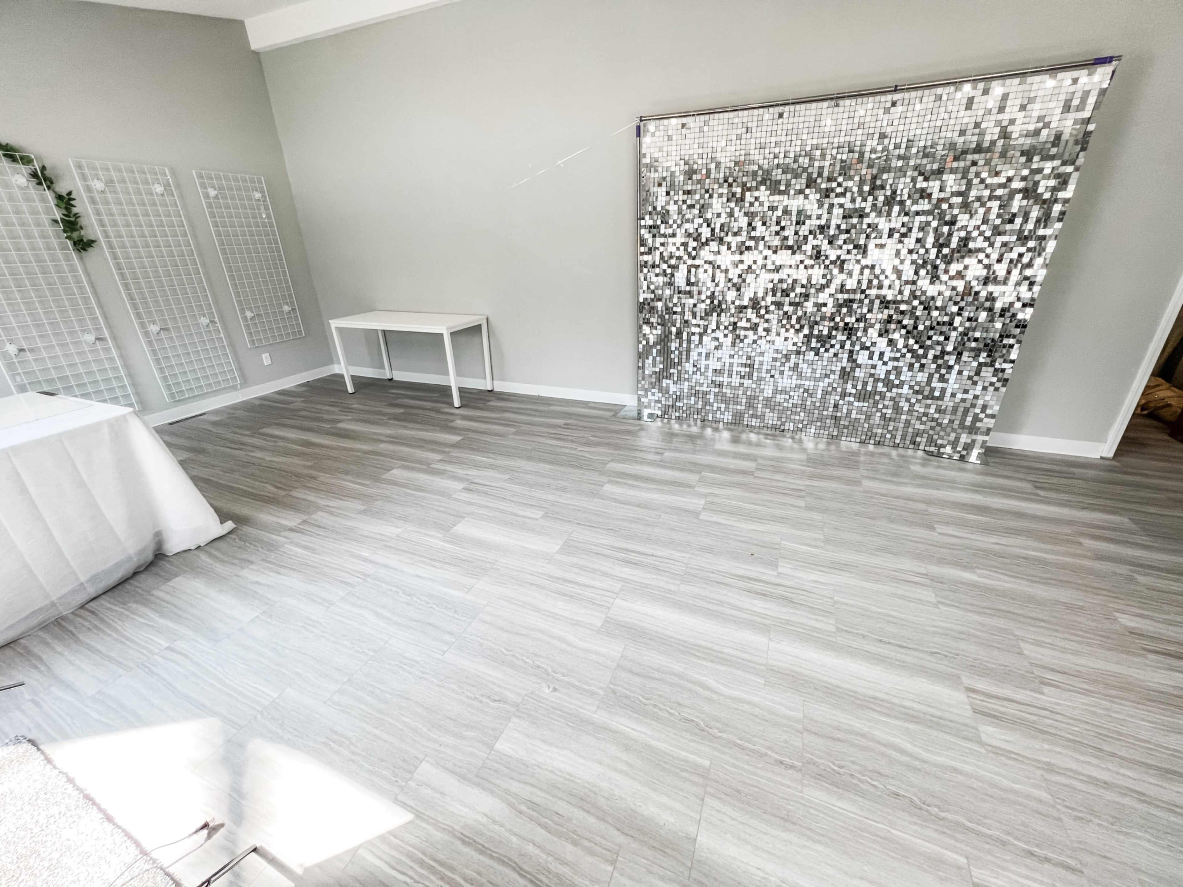 A spacious, well-lit room with gray walls, a light wooden floor, a white table, and a shiny, mirrored wall decoration.