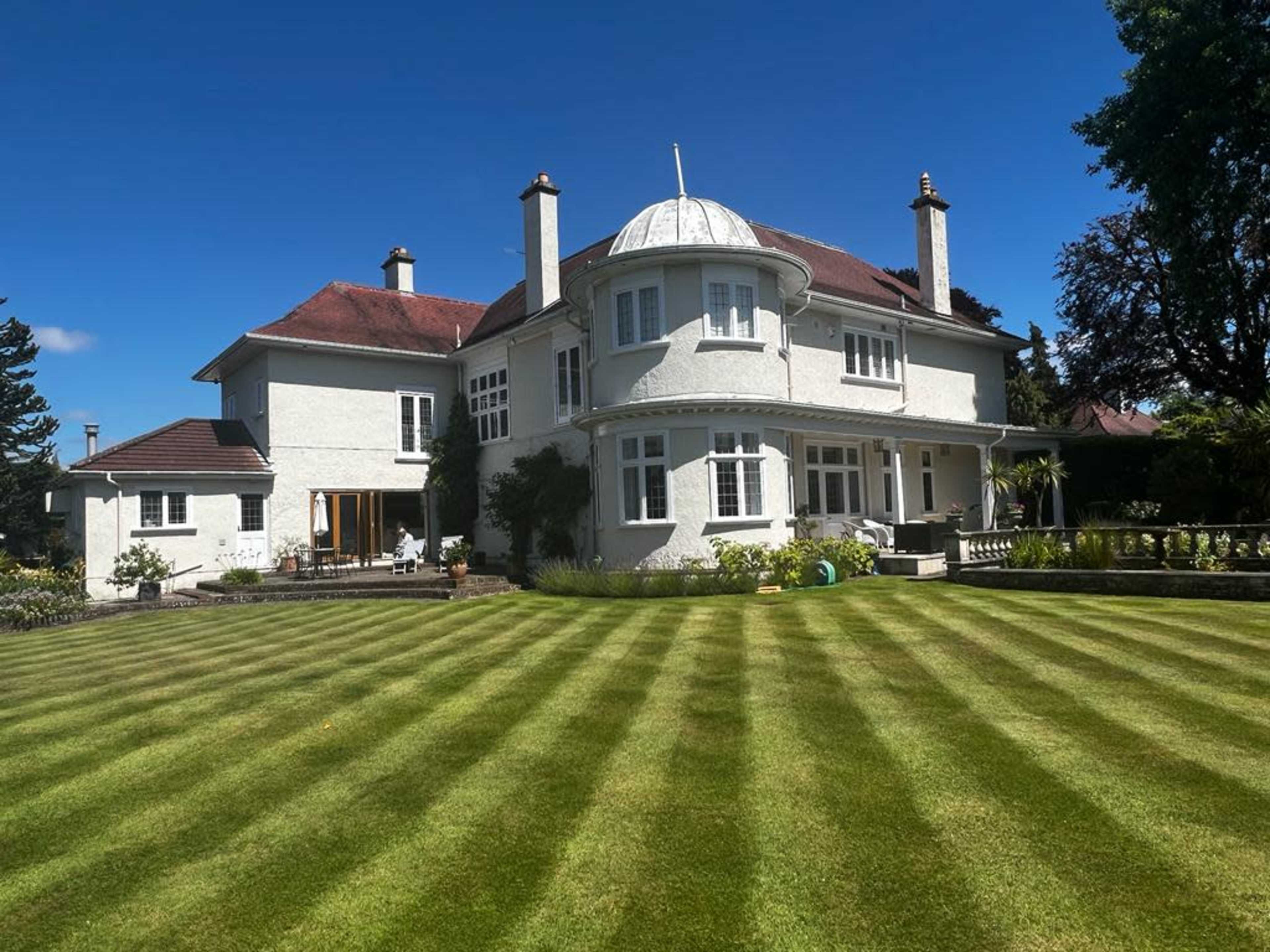 A large, white mansion with a rounded turret and well-maintained lawn features neatly mowed stripes under a clear blue sky.