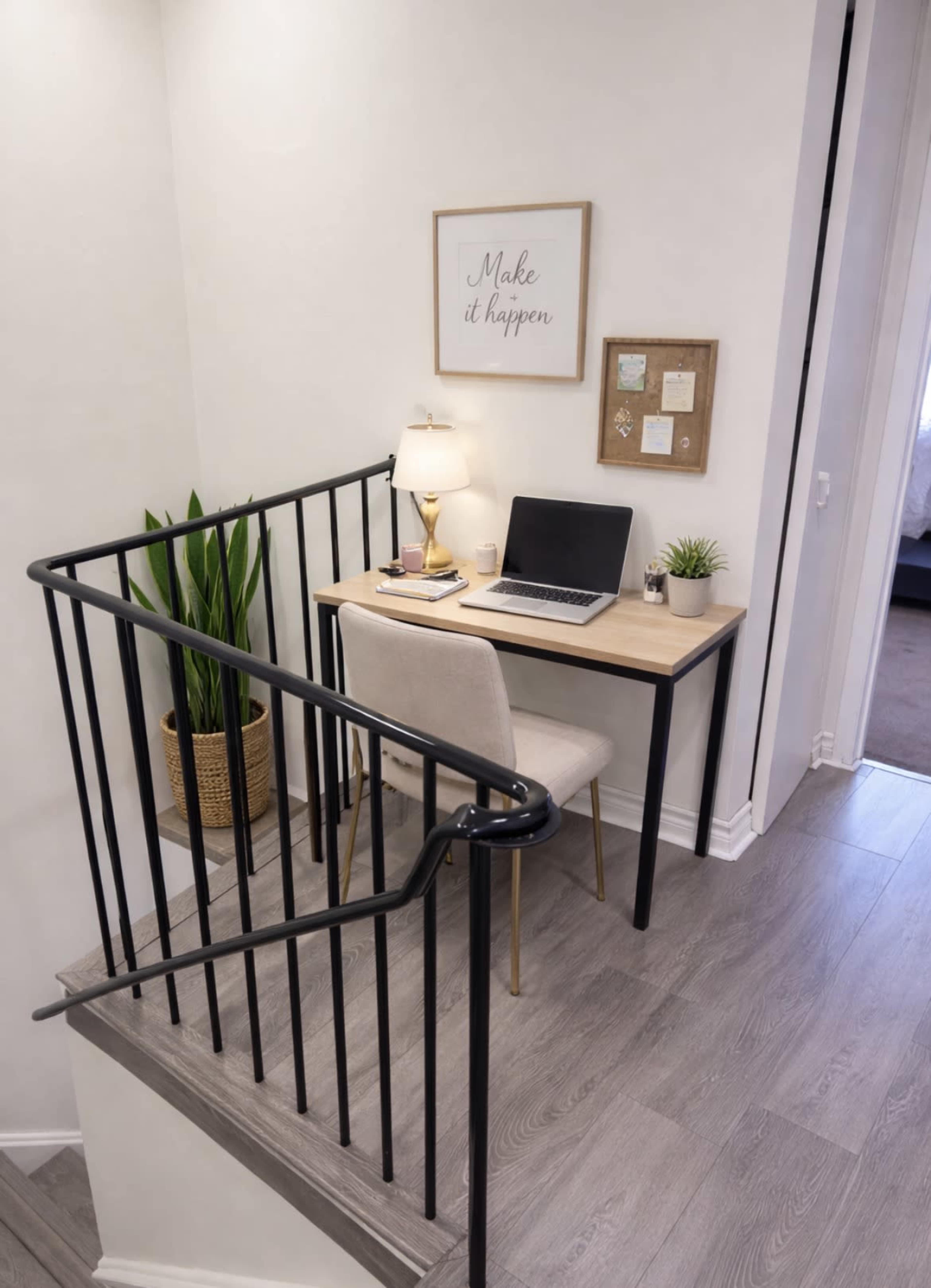 A small workspace with a desk, laptop, and lamp is situated at the bottom of a staircase, accompanied by a potted plant and wall decor.
