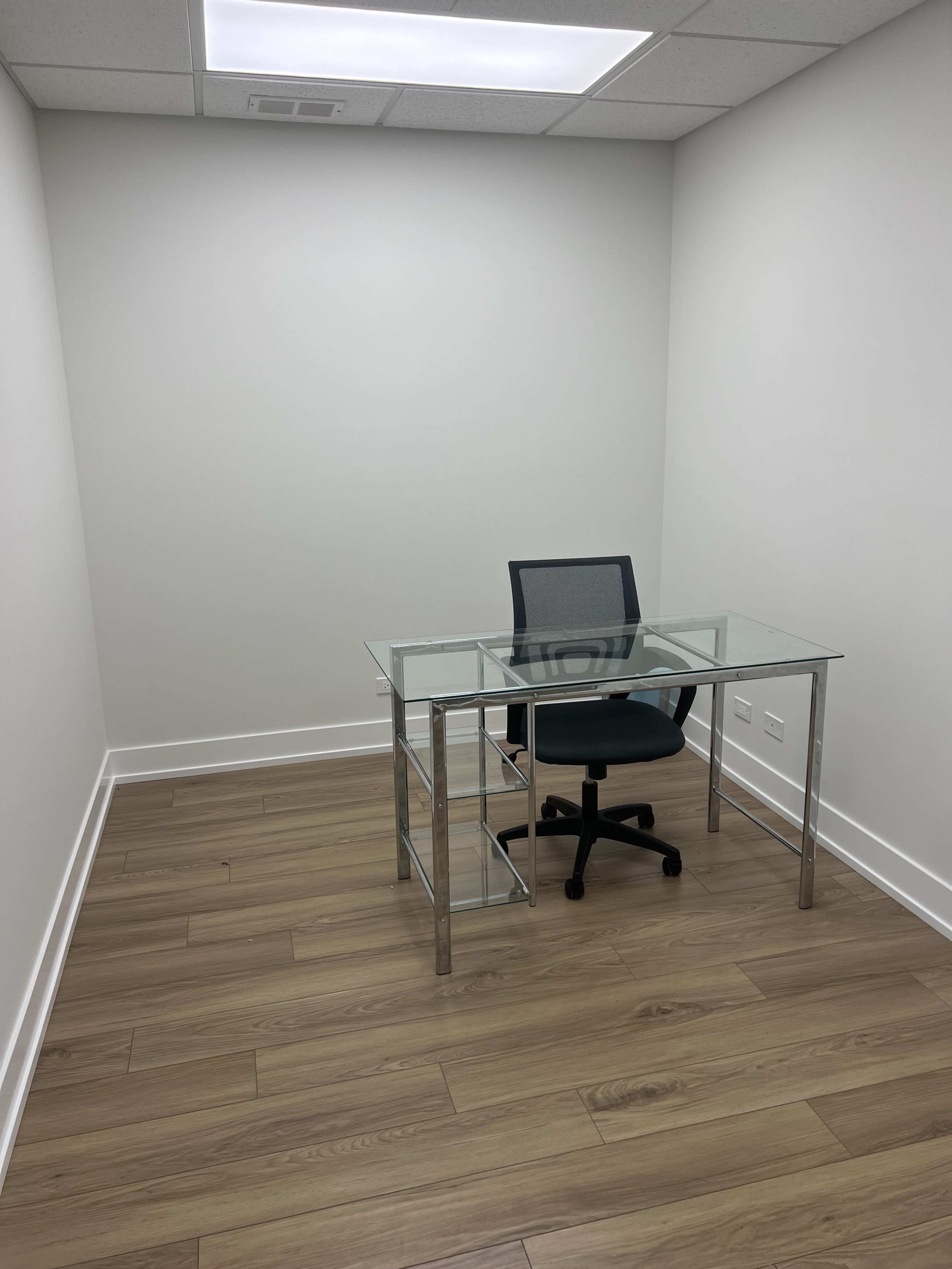 A small, empty office space featuring a glass desk and a swivel chair on a wooden floor.