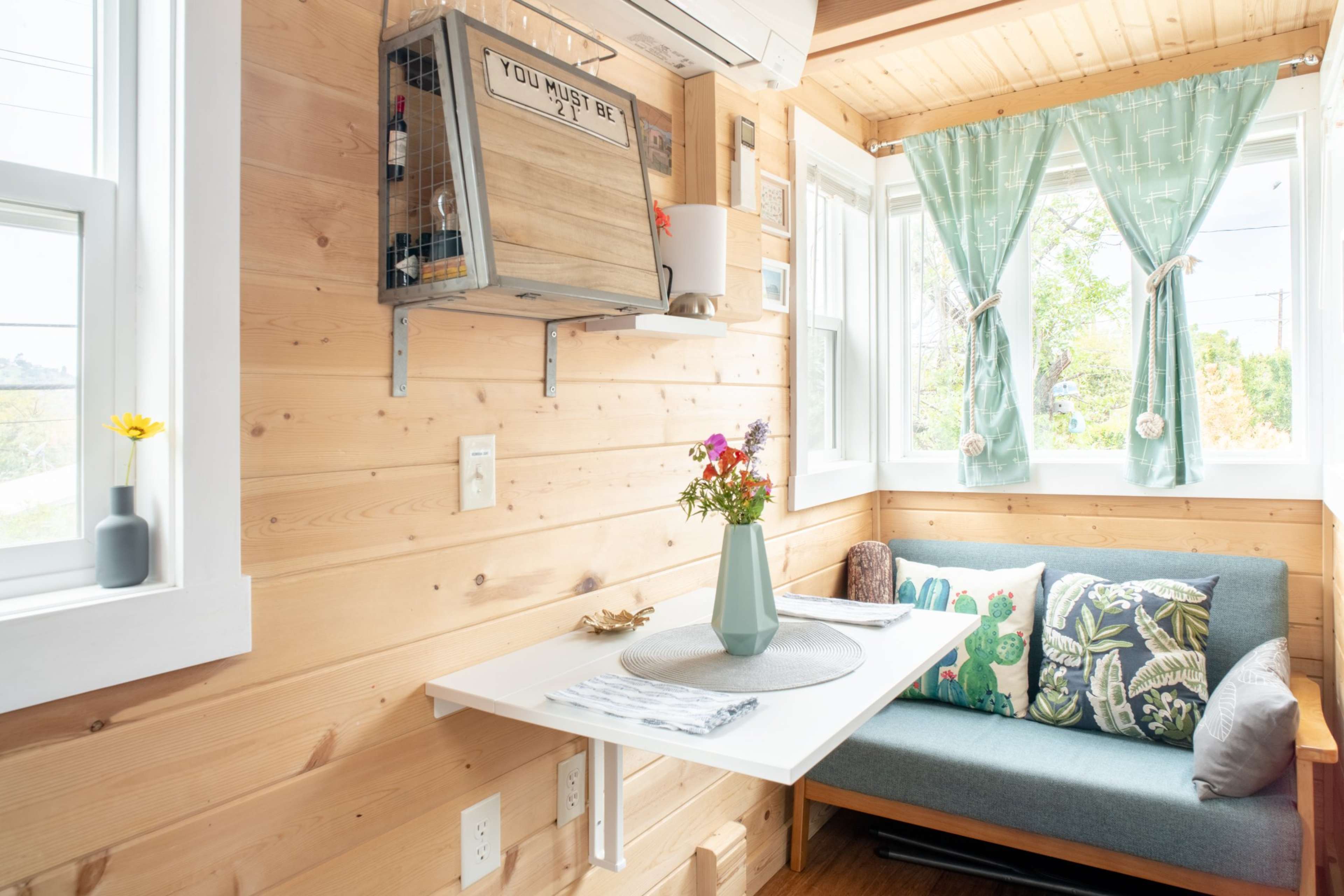 A cozy corner of a small room features a table with a flower vase and a blue couch beside large windows adorned with light green curtains.
