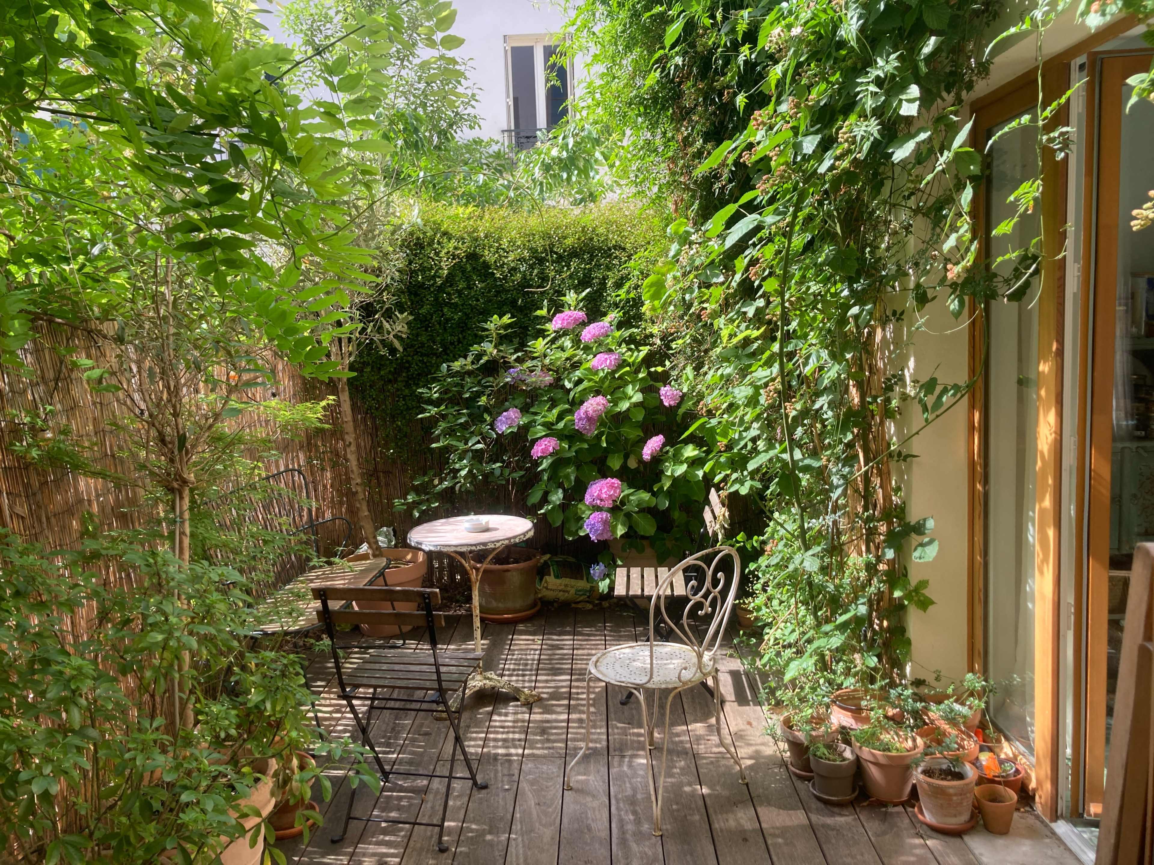 The image shows a small garden patio with wooden flooring, surrounded by lush greenery and blooming hydrangeas, featuring two chairs and a table.