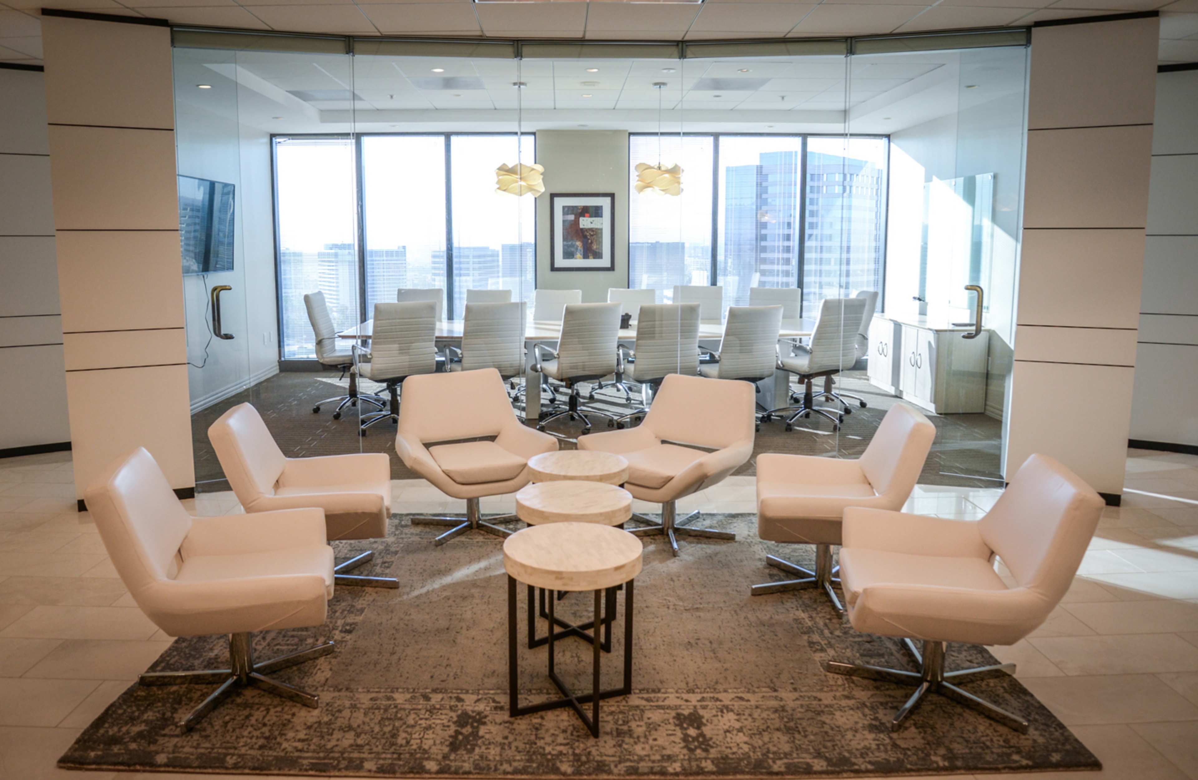 A modern office space featuring a seating area with white chairs arranged around two small tables, next to a conference room visible through glass walls.
