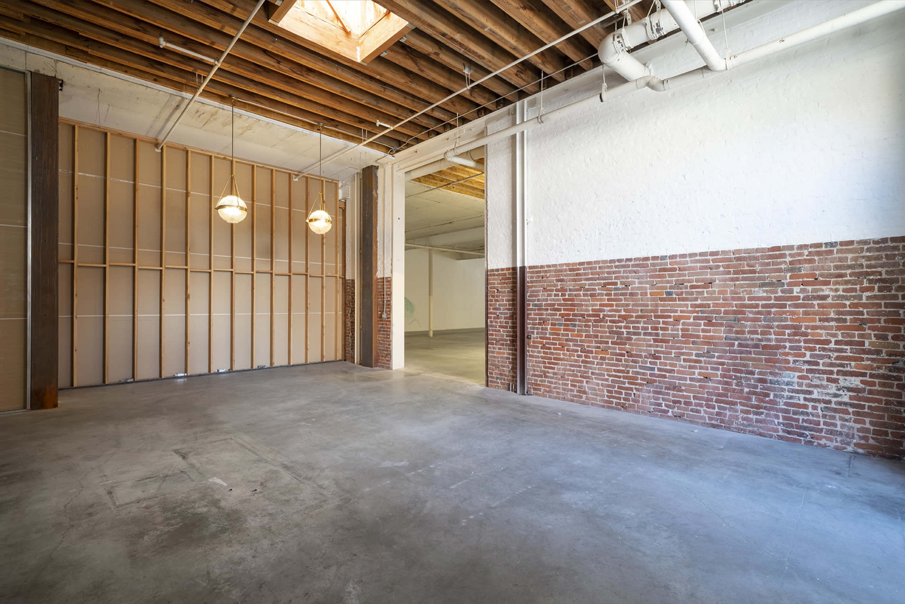 An empty industrial space with exposed brick walls and wooden beams overhead, featuring two hanging light fixtures.