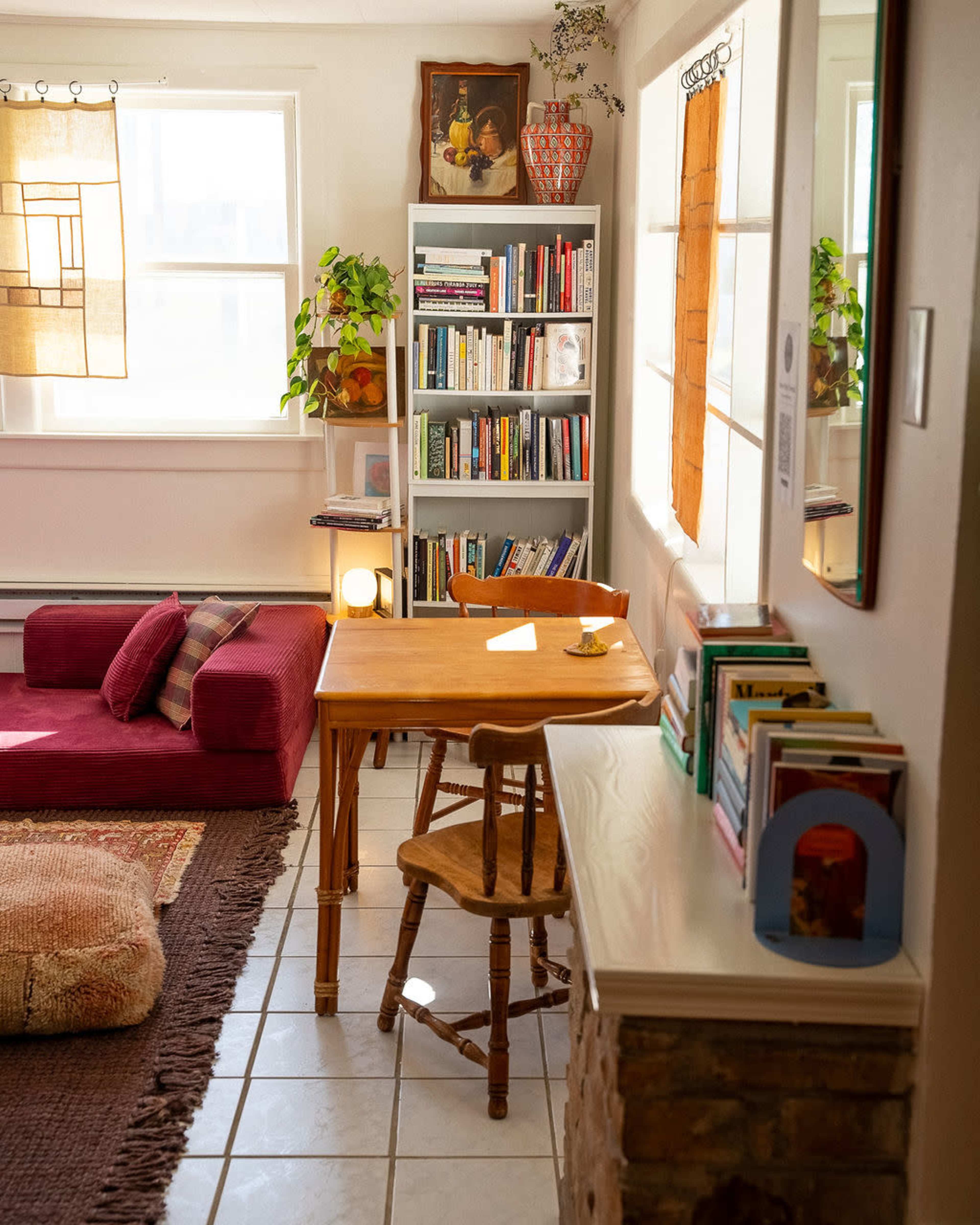 The image shows a cozy living space featuring a small wooden table, a bookshelf filled with books, a seating area with cushions, and natural light coming through the windows.