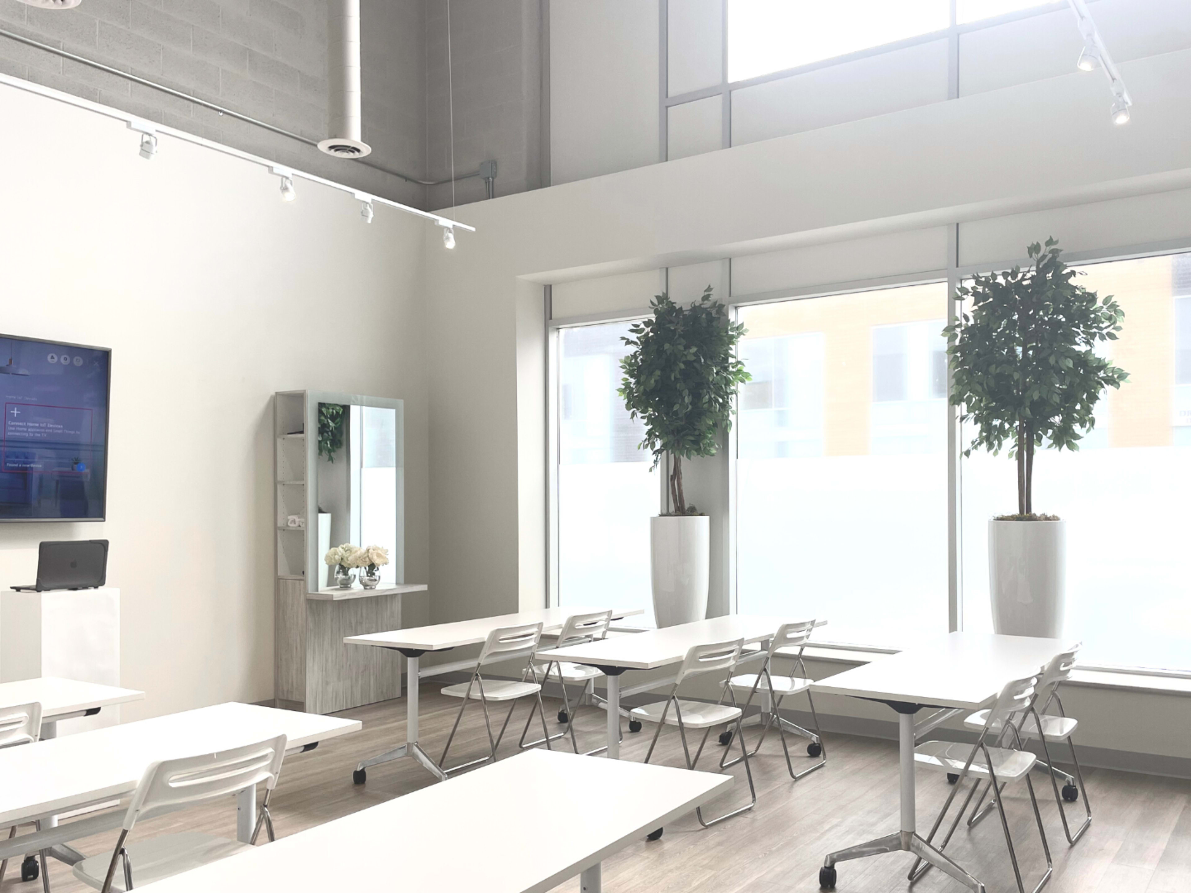 A modern classroom with several white tables and chairs, large windows, and potted plants in tall white planters.