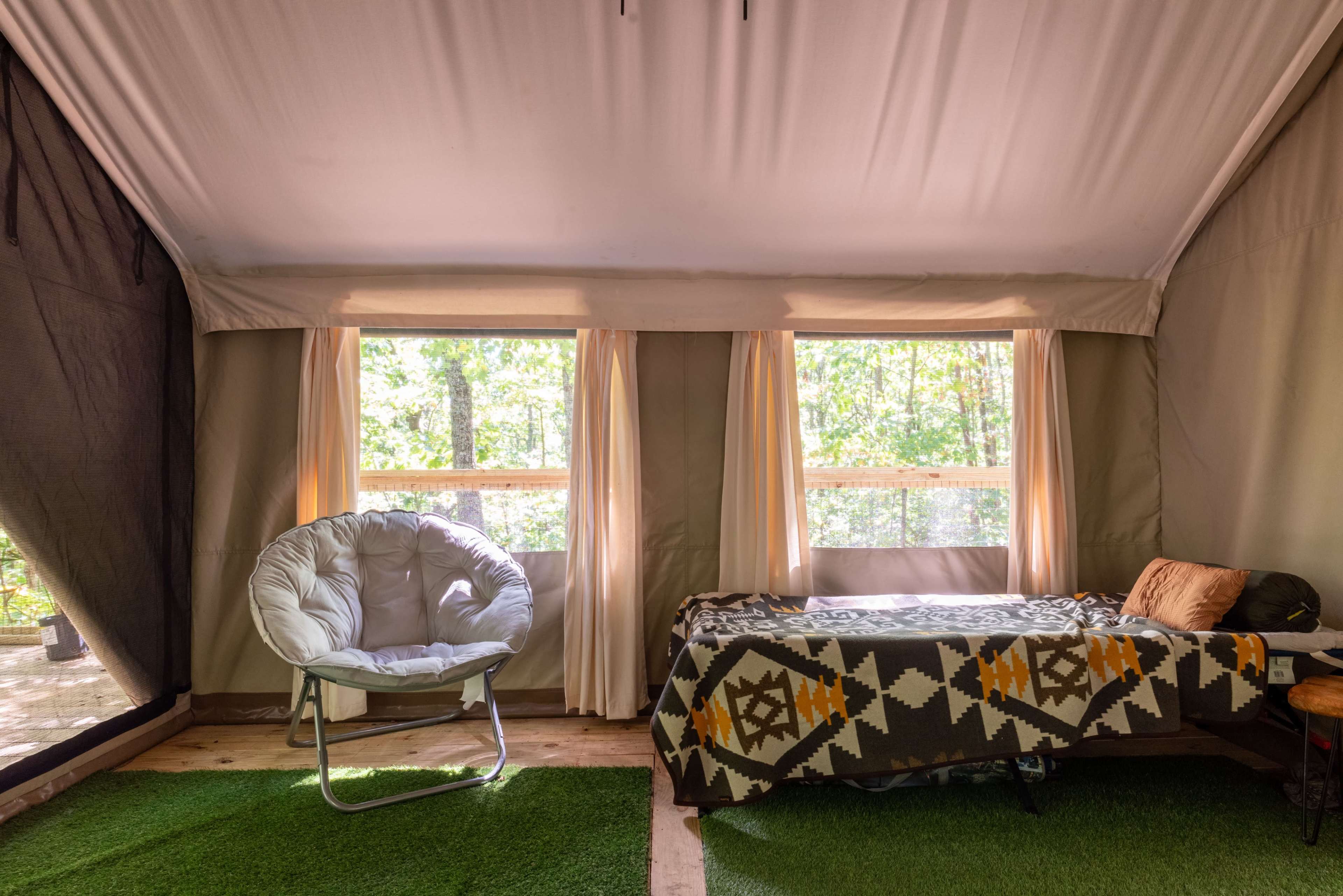 The interior of a canvas tent features a chair and a bed, with large windows allowing natural light and views of greenery outside.