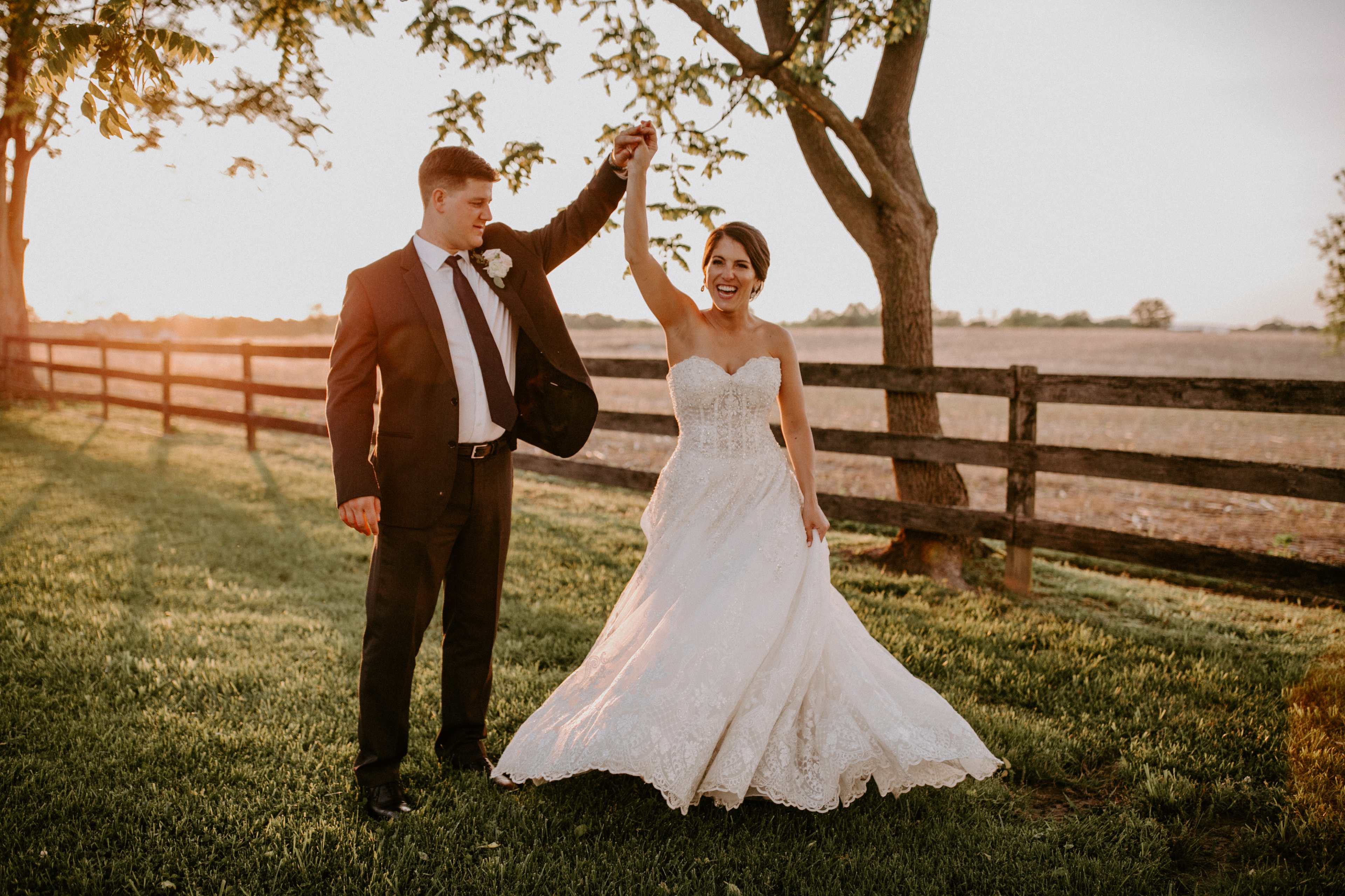 A man in a suit lifts the arm of a woman in a wedding dress while standing on a grassy area near a wooden fence at sunset.
