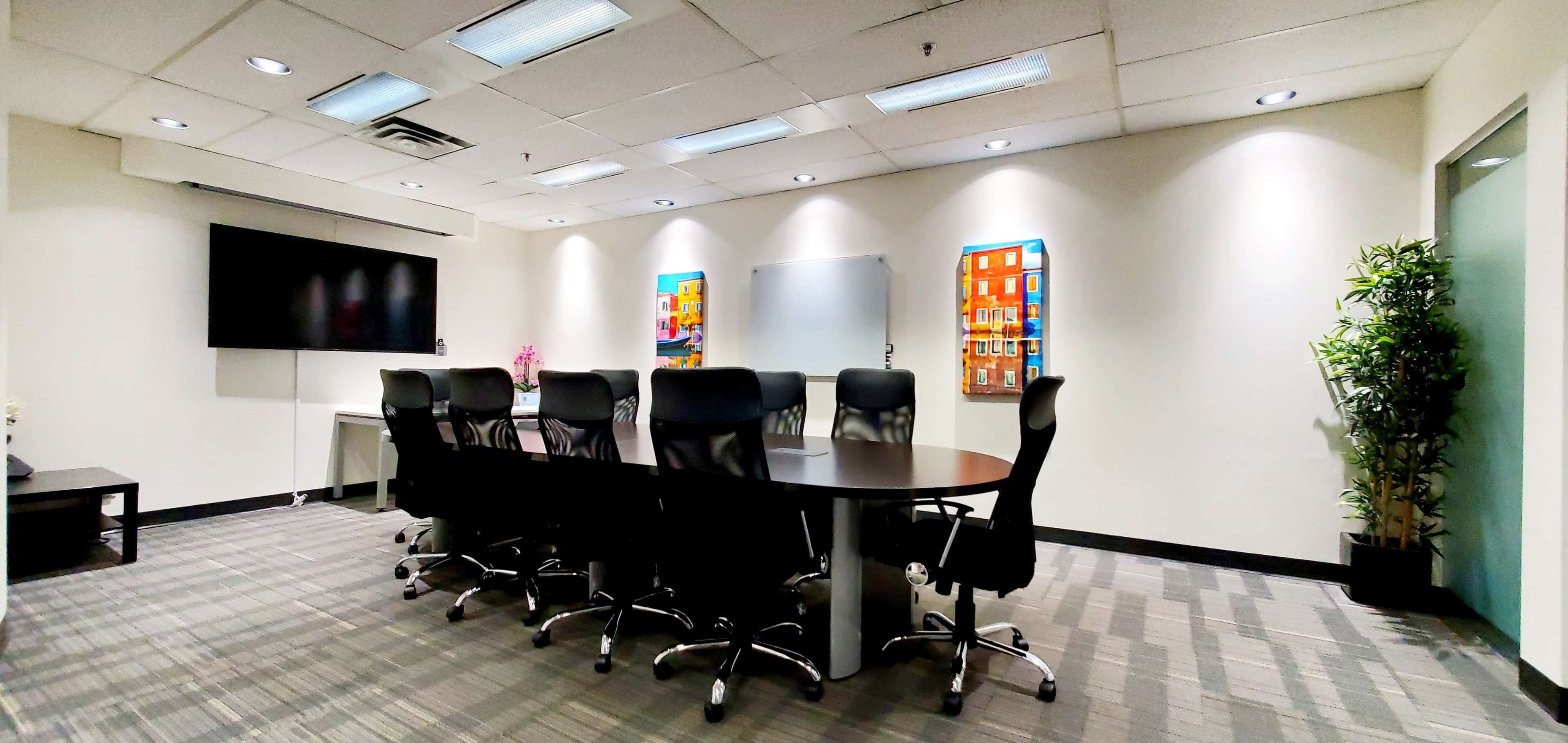 A conference room features a large oval table surrounded by black ergonomic chairs, a wall-mounted television, and two framed artworks.
