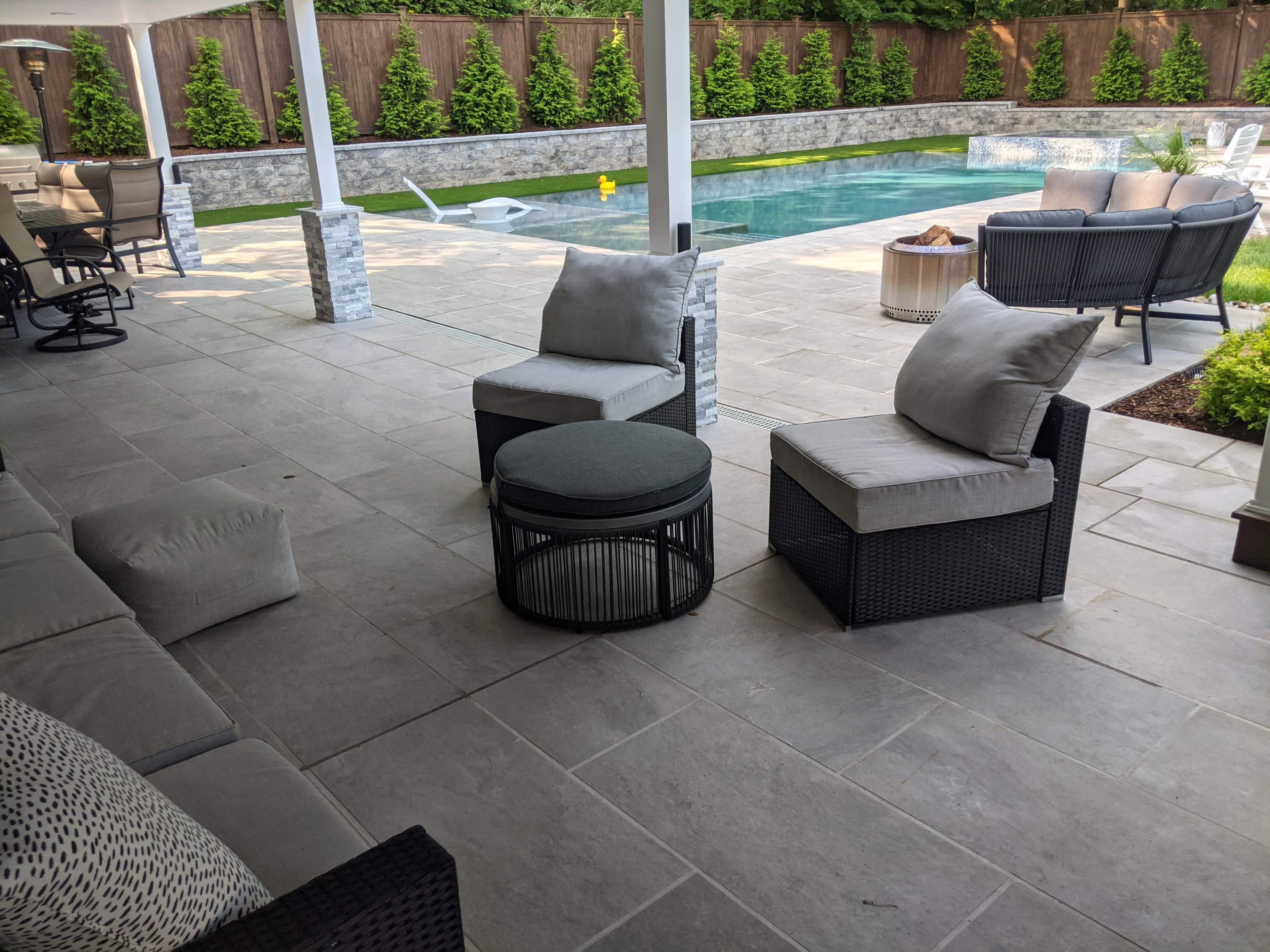A patio area featuring two cushioned chairs and a round table, overlooking a swimming pool surrounded by landscaped greenery.