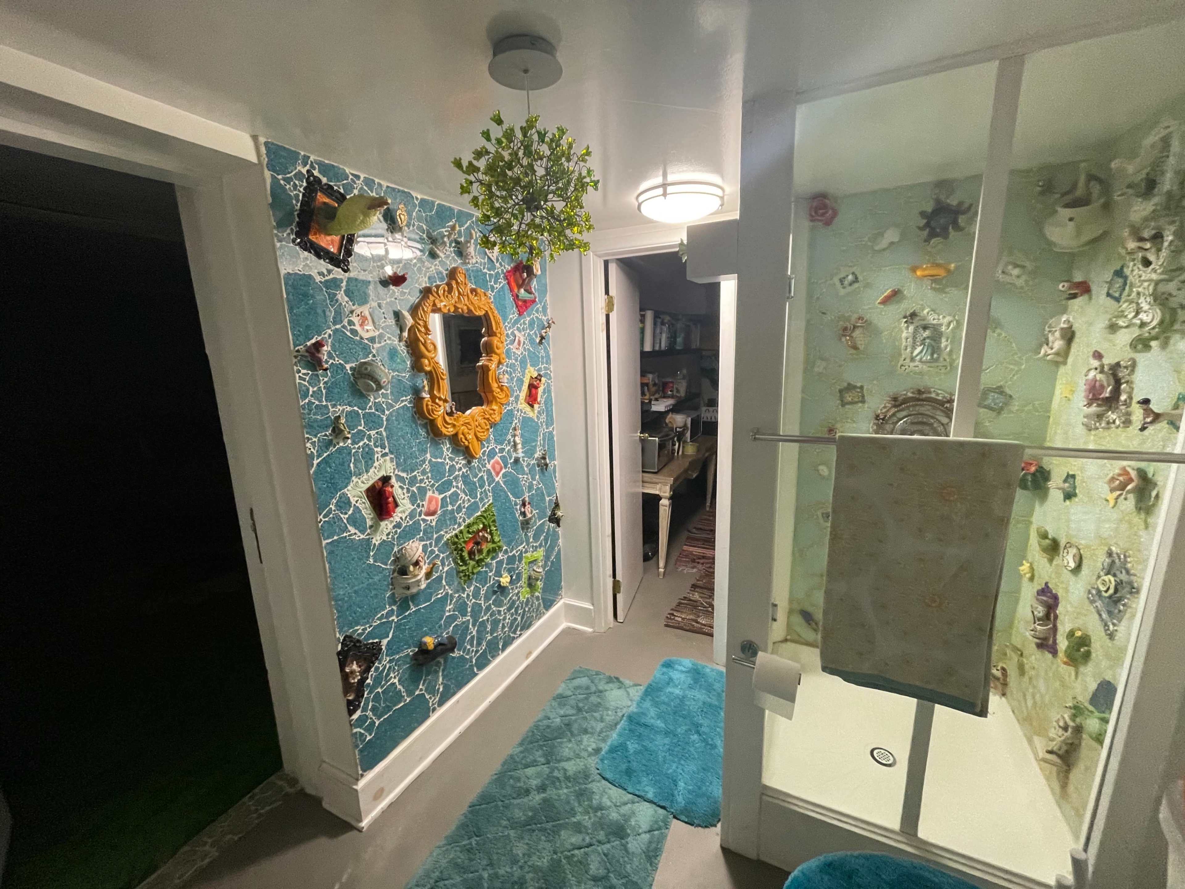 A bathroom with a wall decorated in a mosaic style featuring various objects, a mirror, and a shower area behind transparent glass.