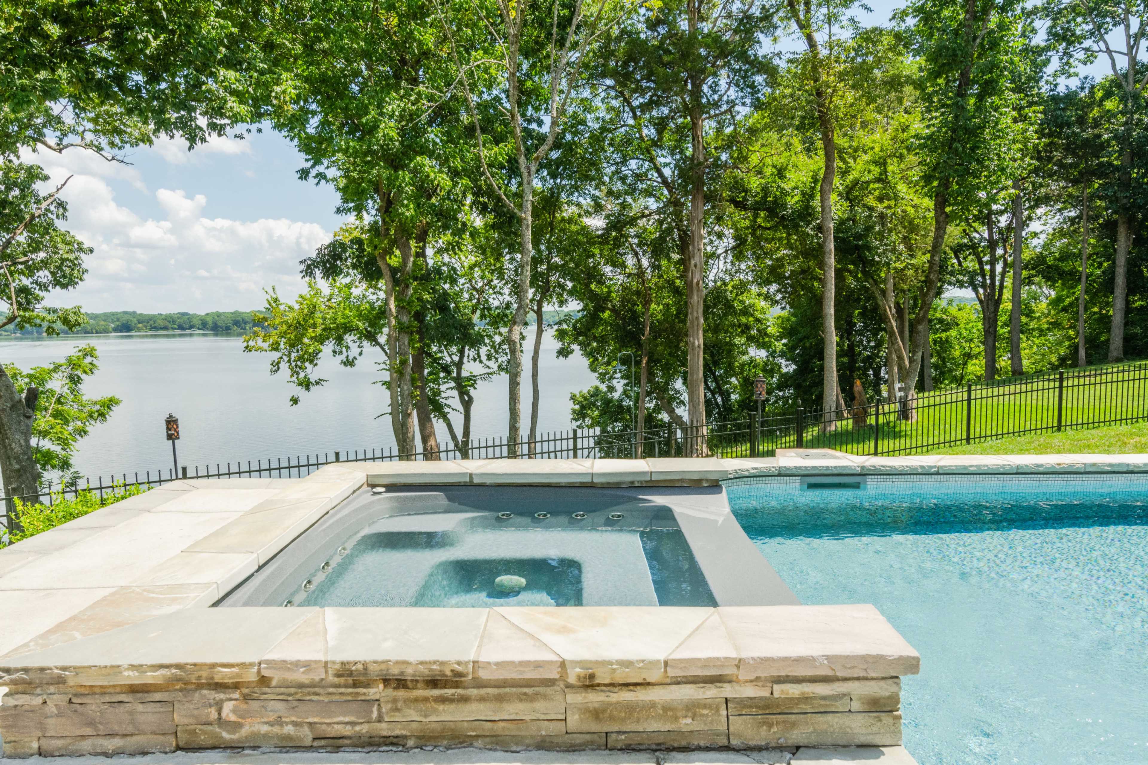 A swimming pool with a hot tub overlooking a lake, surrounded by trees and green grass.