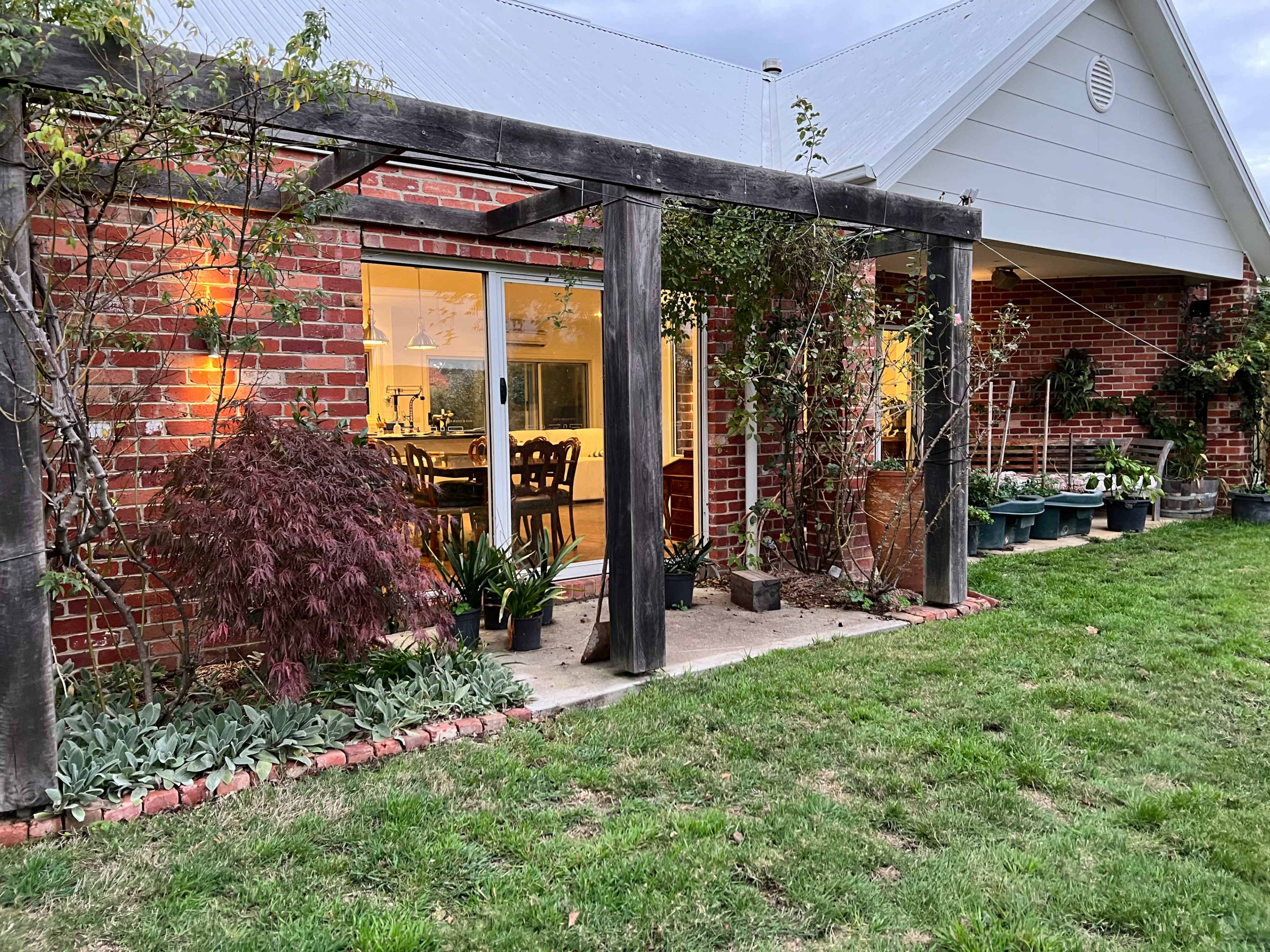 A brick house with a wooden pergola covered in vines, leading to a brightly lit interior with a dining area visible through large sliding glass doors.