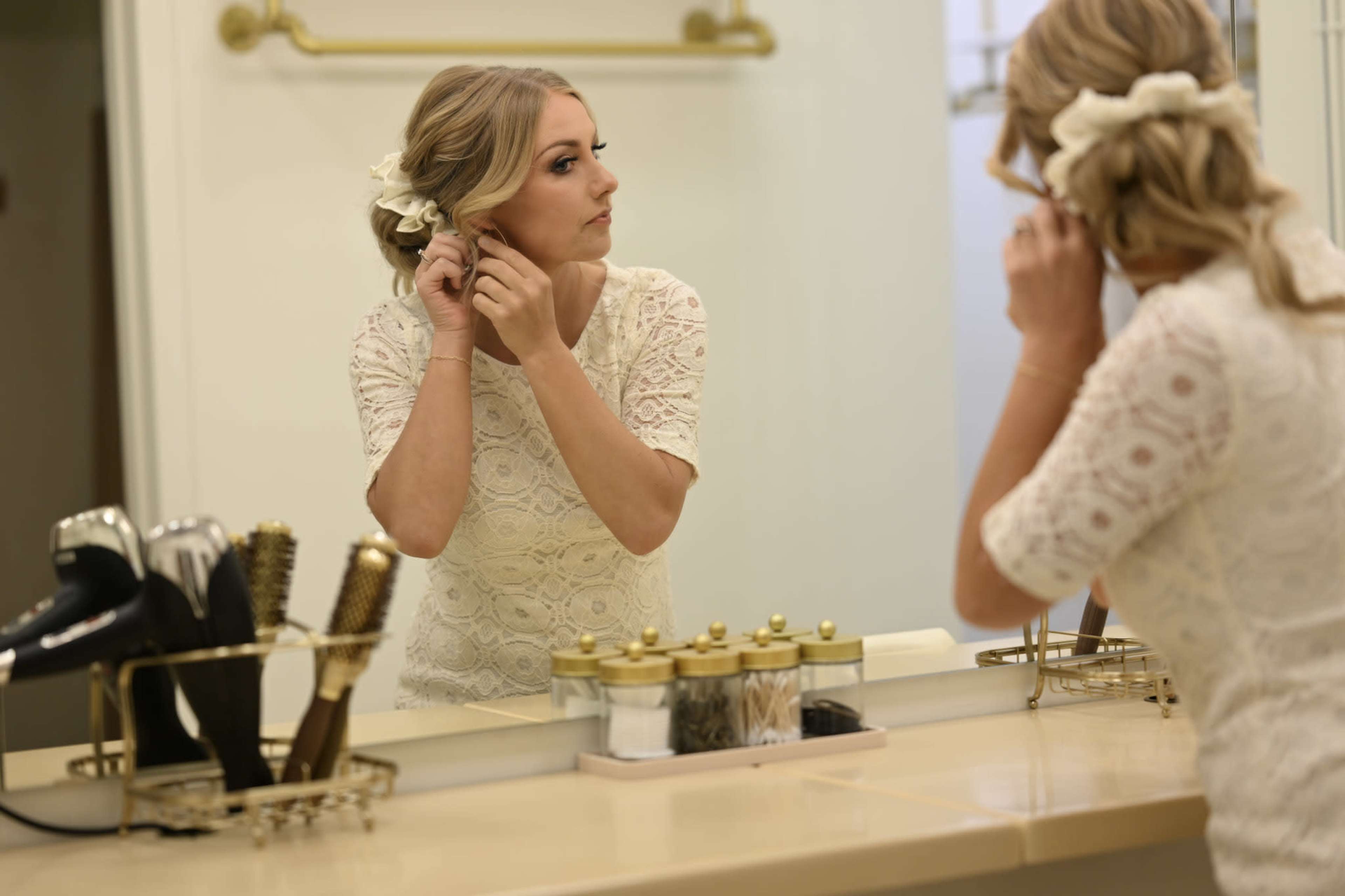 A woman in a lace dress adjusts her hair while looking in a mirror in a bathroom.