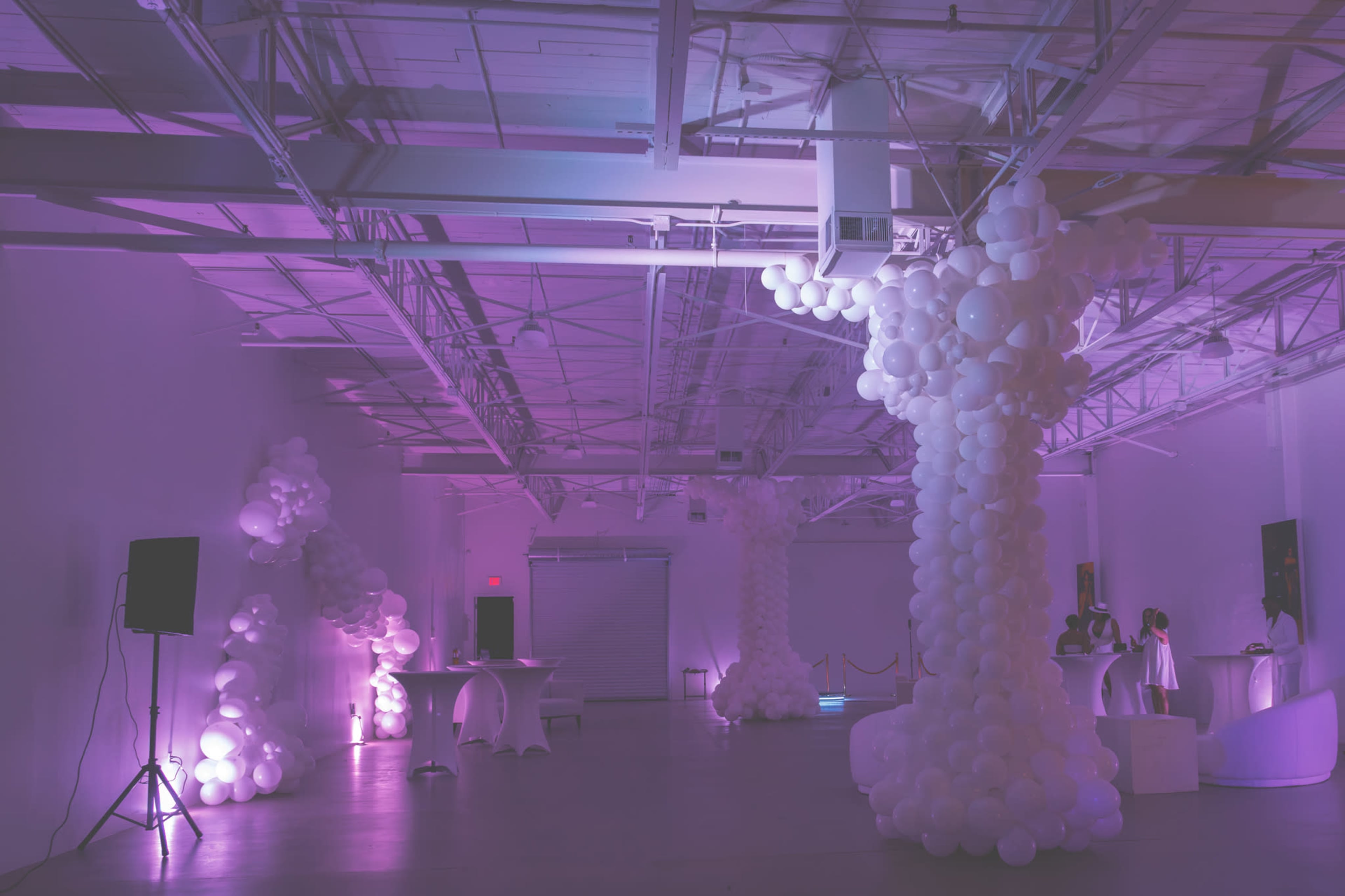 A spacious event venue decorated with large white balloon installations and dim purple lighting.