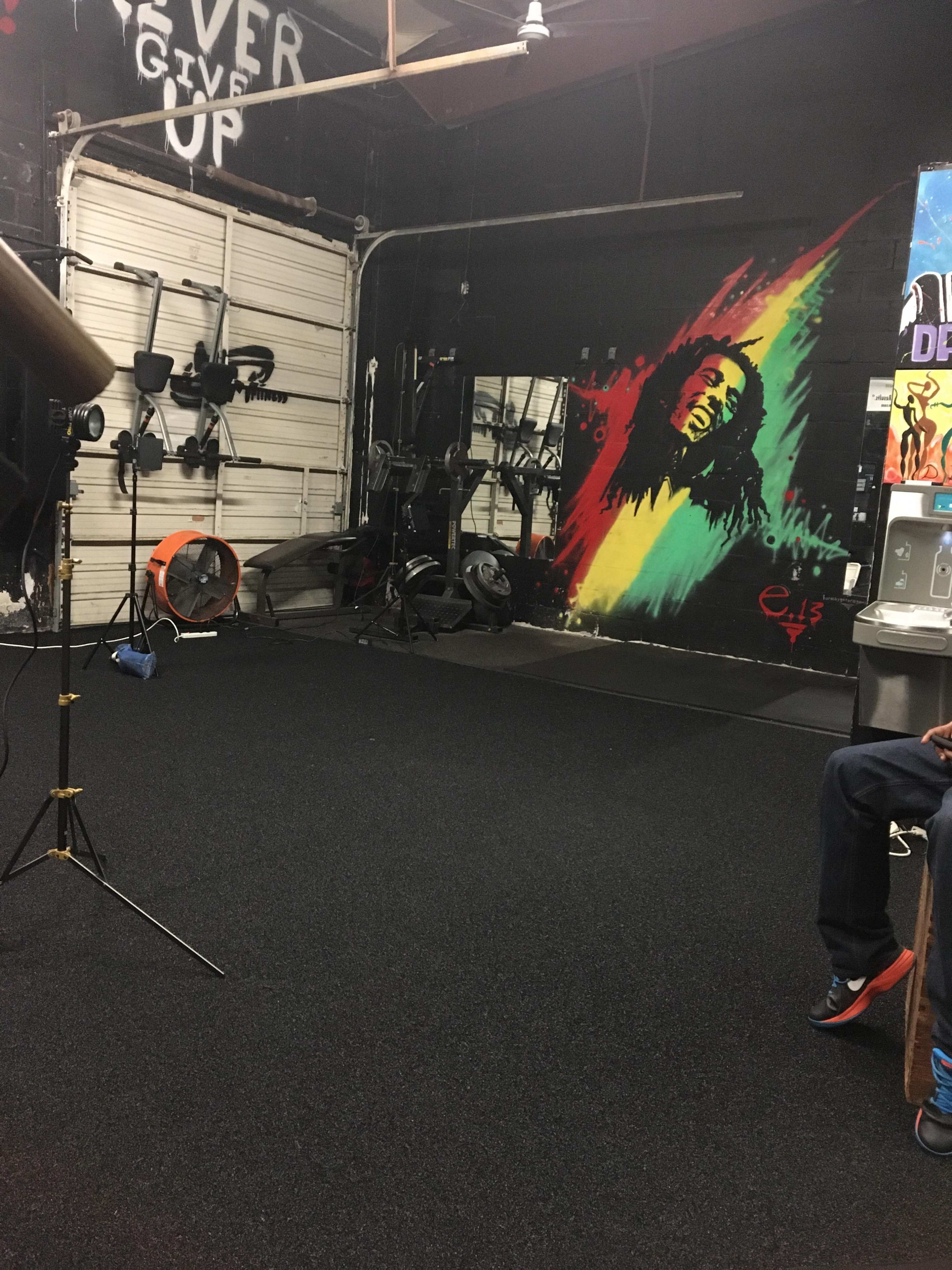 A gym with workout equipment, a colorful mural of a reggae artist on the wall, and motivational graffiti.