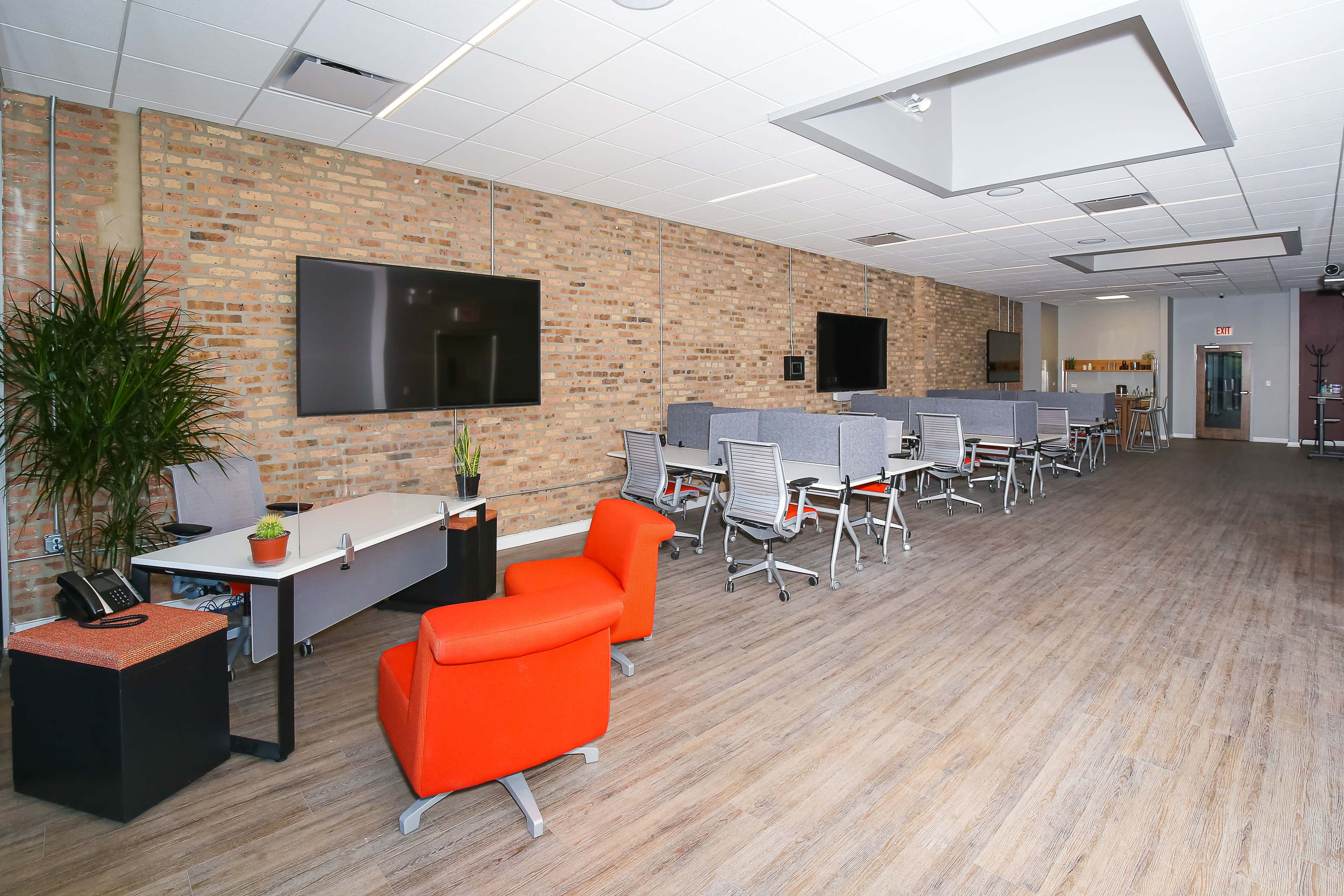 A spacious office area with a mix of workstations, a seating area, and two large television screens mounted on a brick wall.