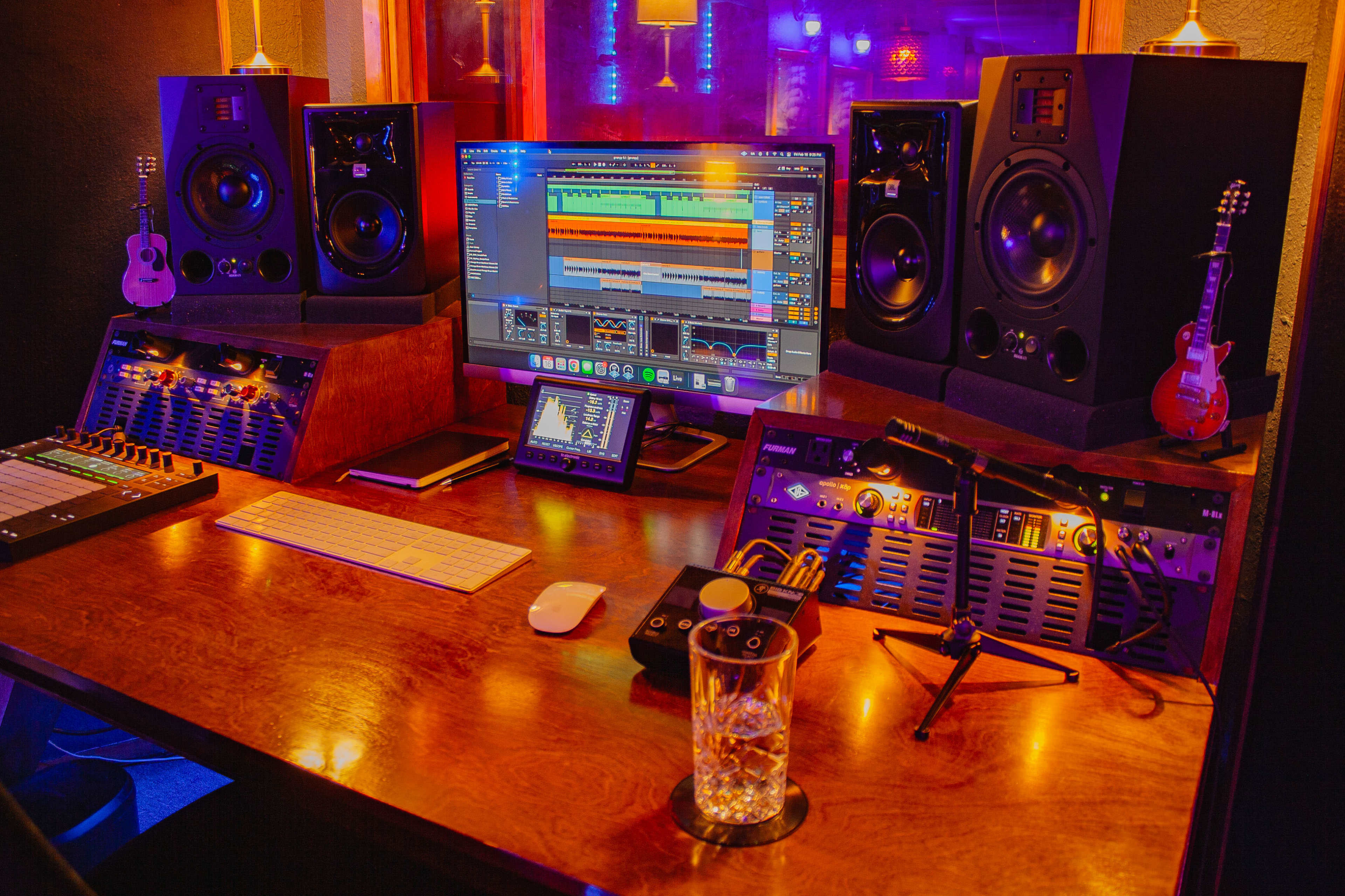 The image shows a music production studio with a wooden desk housing computer screens, audio equipment, and small guitars, alongside a glass of water.