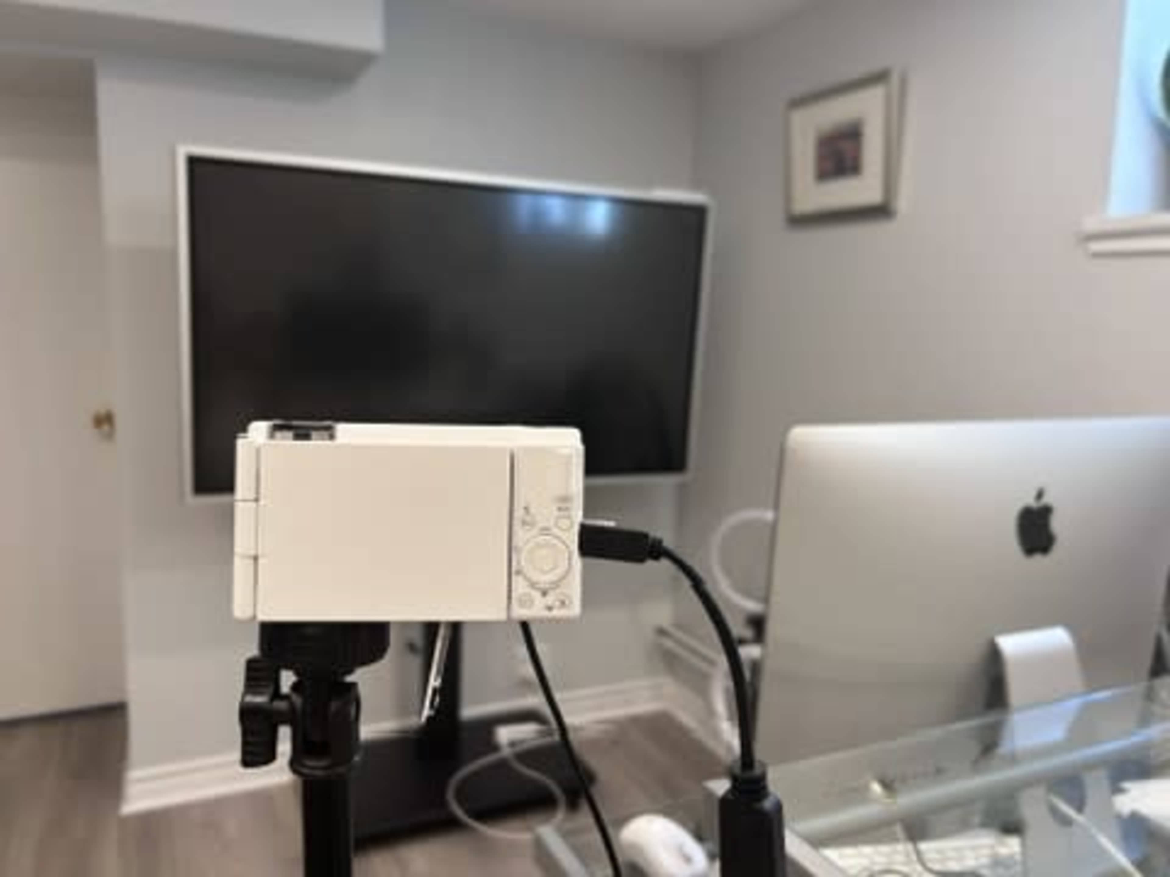 A white camera is mounted on a tripod and connected to a monitor in a modern office setting.