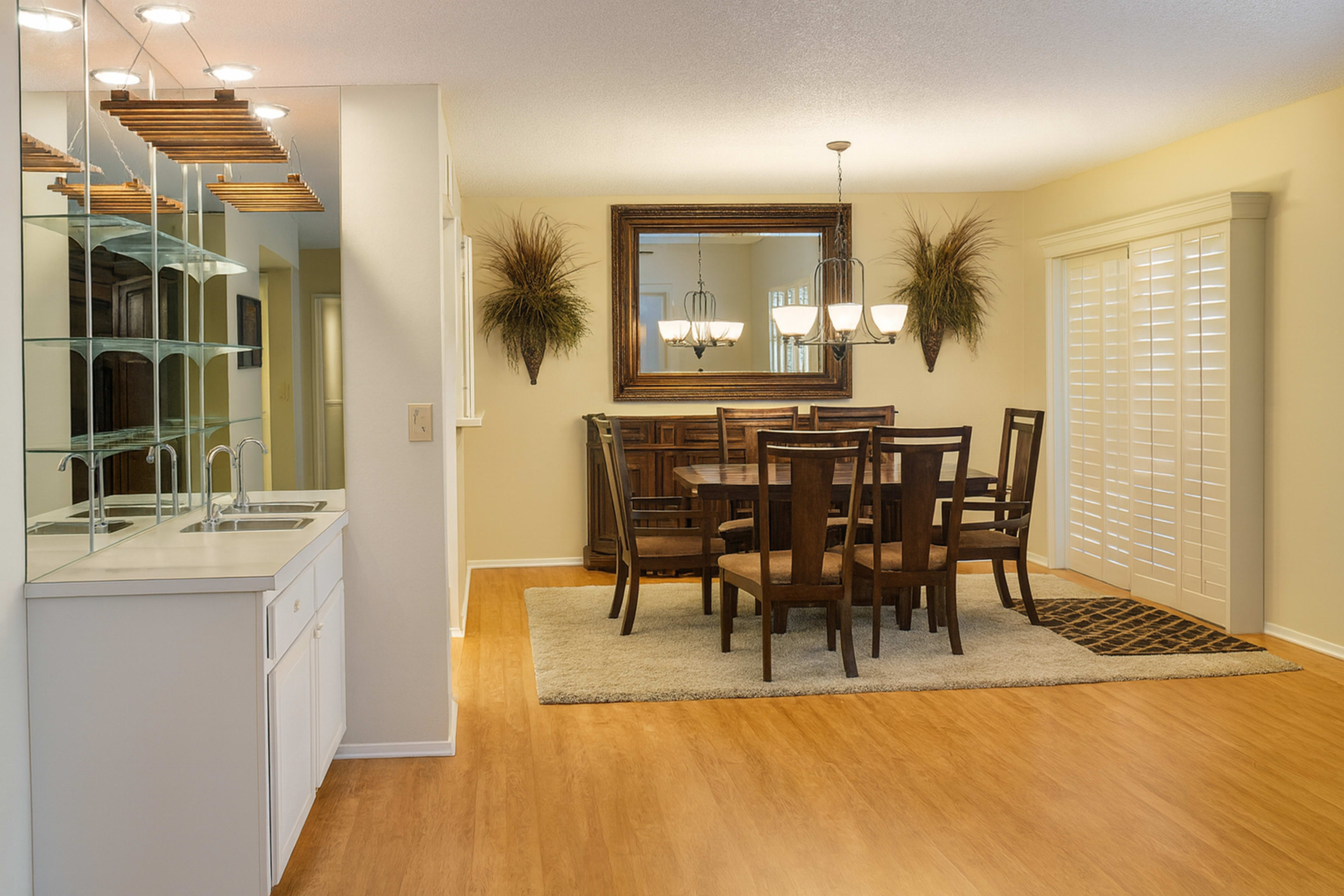 The image shows a well-lit dining area featuring a wooden table surrounded by chairs, with a sideboard and a large mirror on the wall.