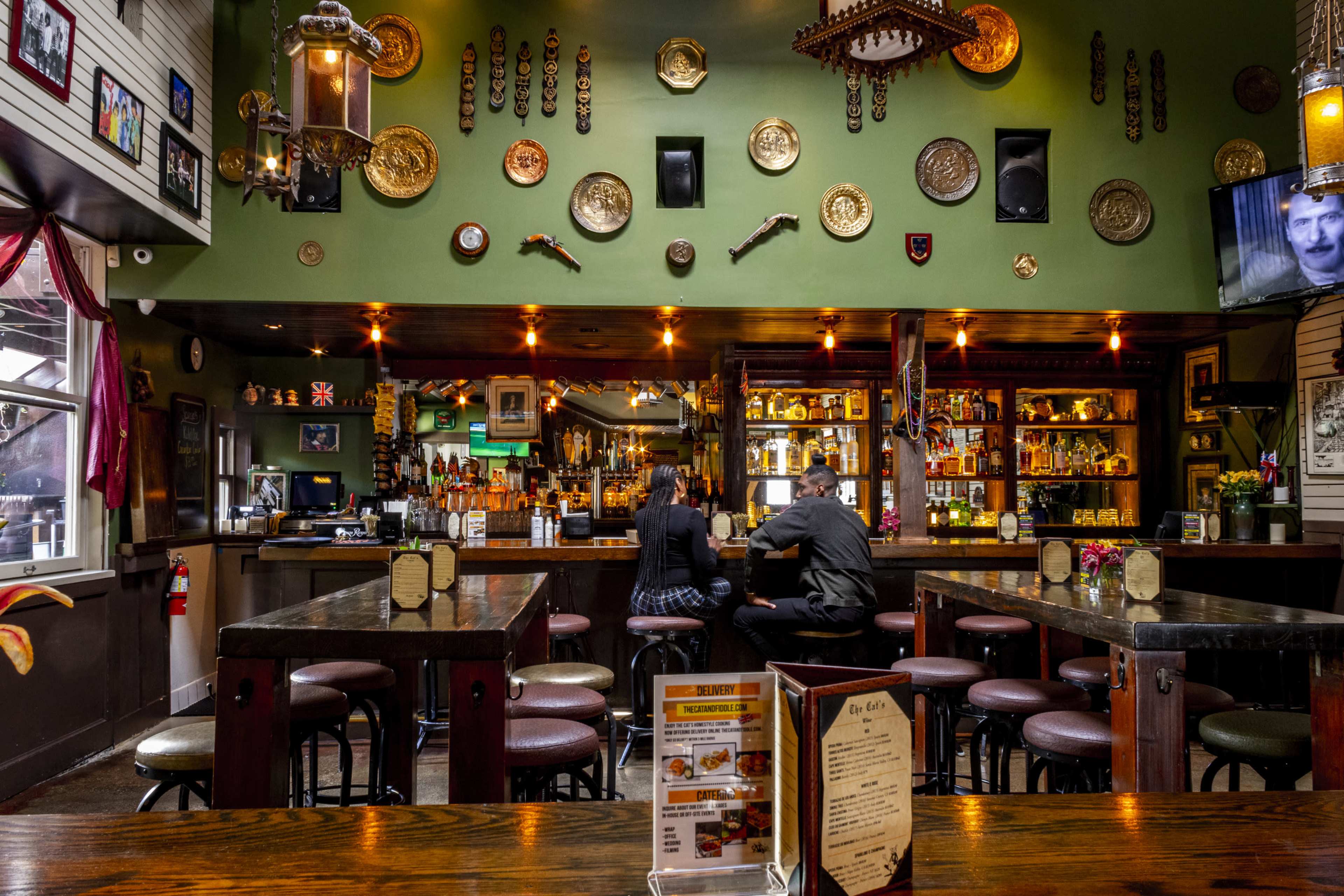 Two patrons sit at the bar in a pub decorated with various plates and artifacts, while shelves filled with bottles are visible behind them.