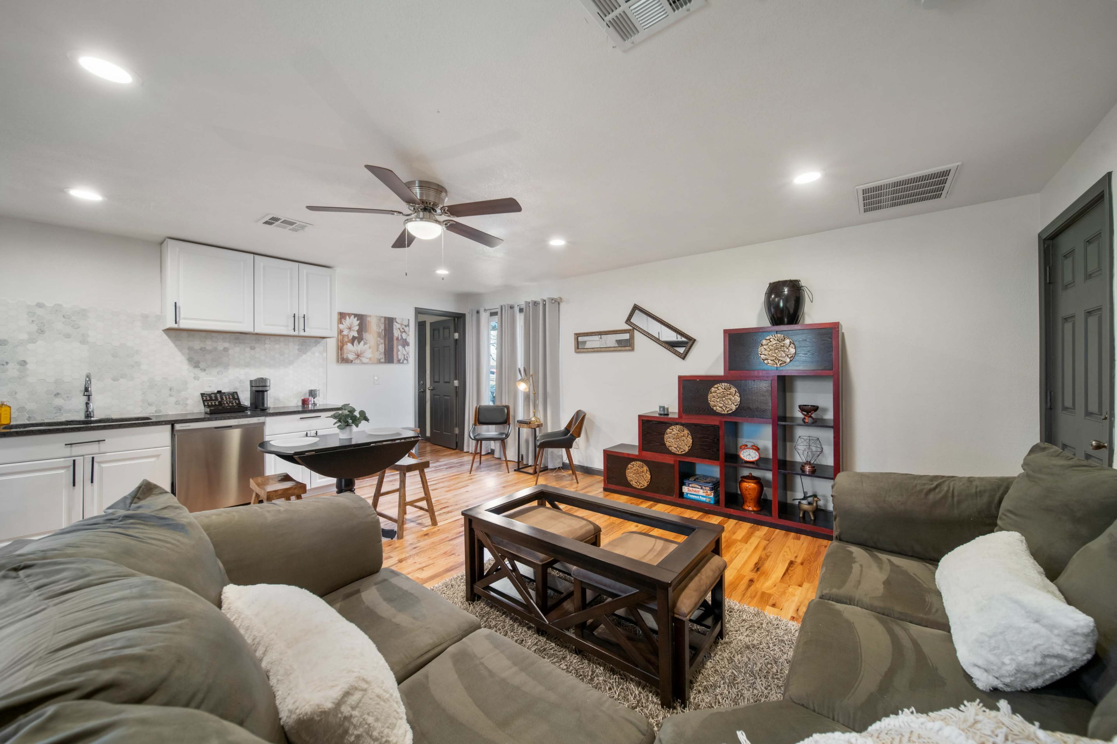 The living area features a sectional sofa, a wooden coffee table, a dining table, and open shelving against a light-colored wall with a ceiling fan above.