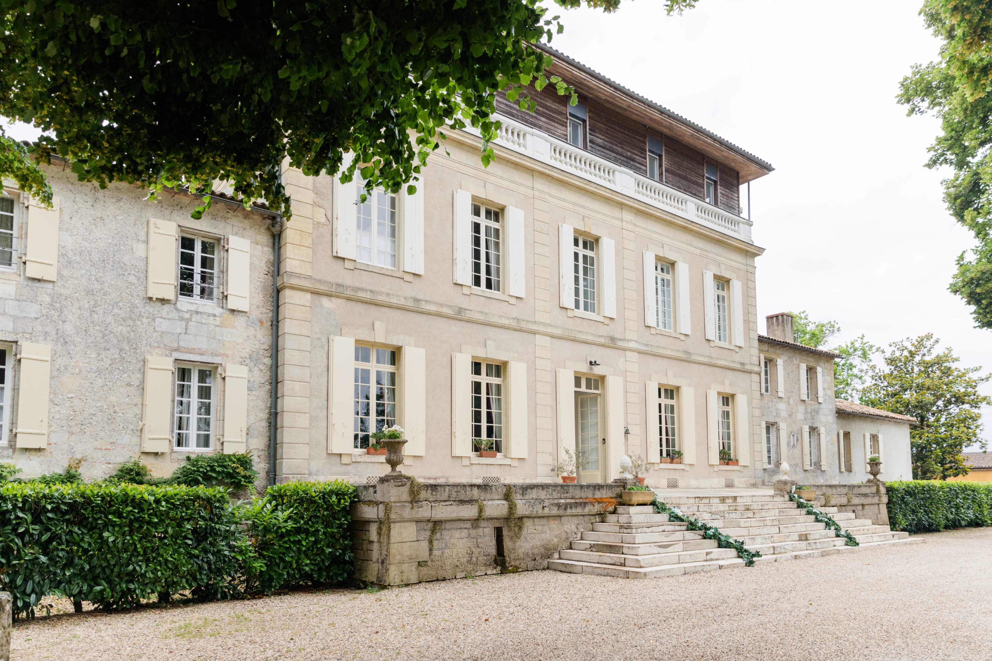 The image shows a large, three-story stone house with a balcony, surrounded by greenery and a gravel pathway leading to its entrance.