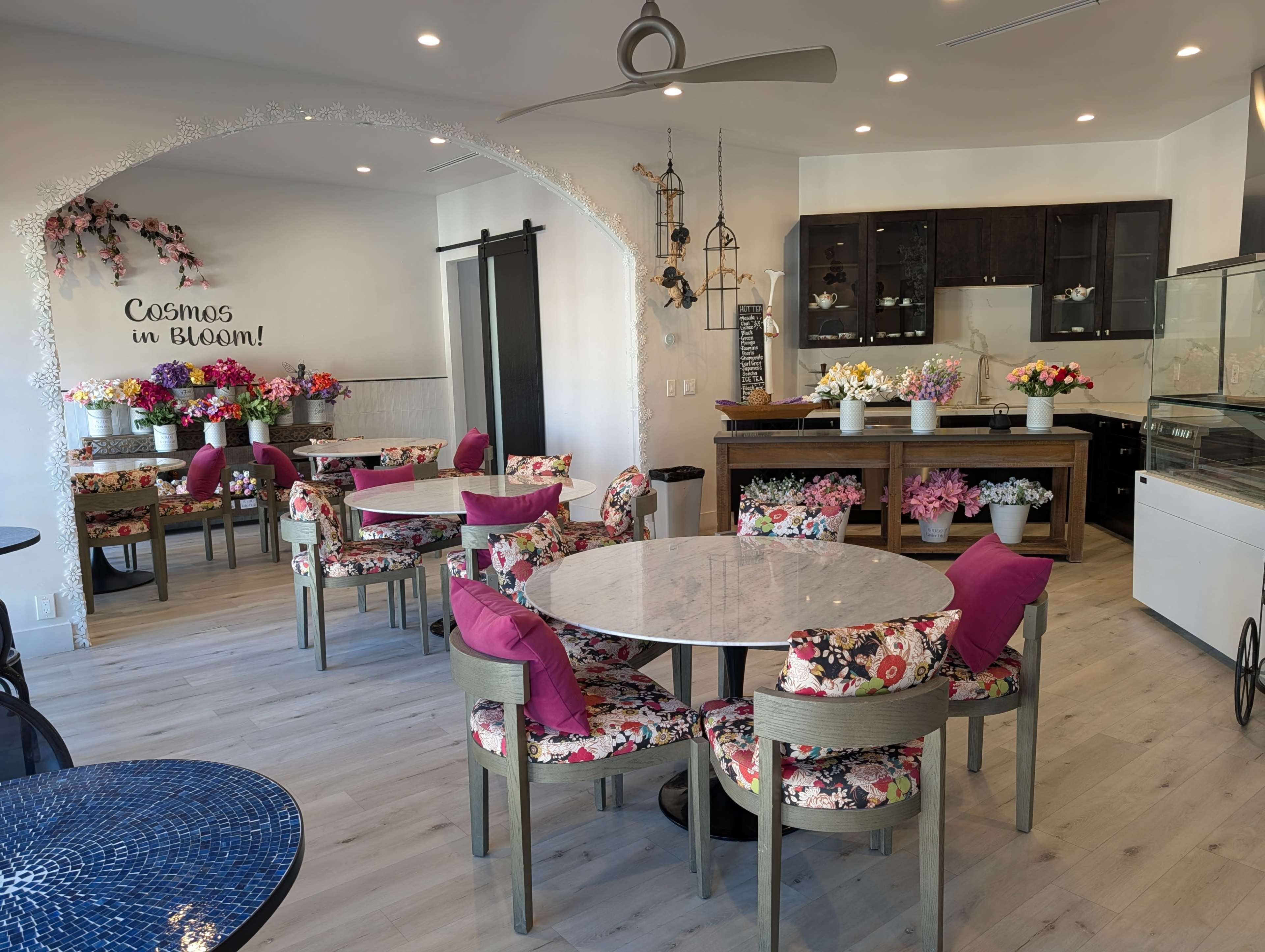 The image shows a bright café interior with floral-patterned chairs, marble tables, and a counter adorned with colorful flower arrangements.