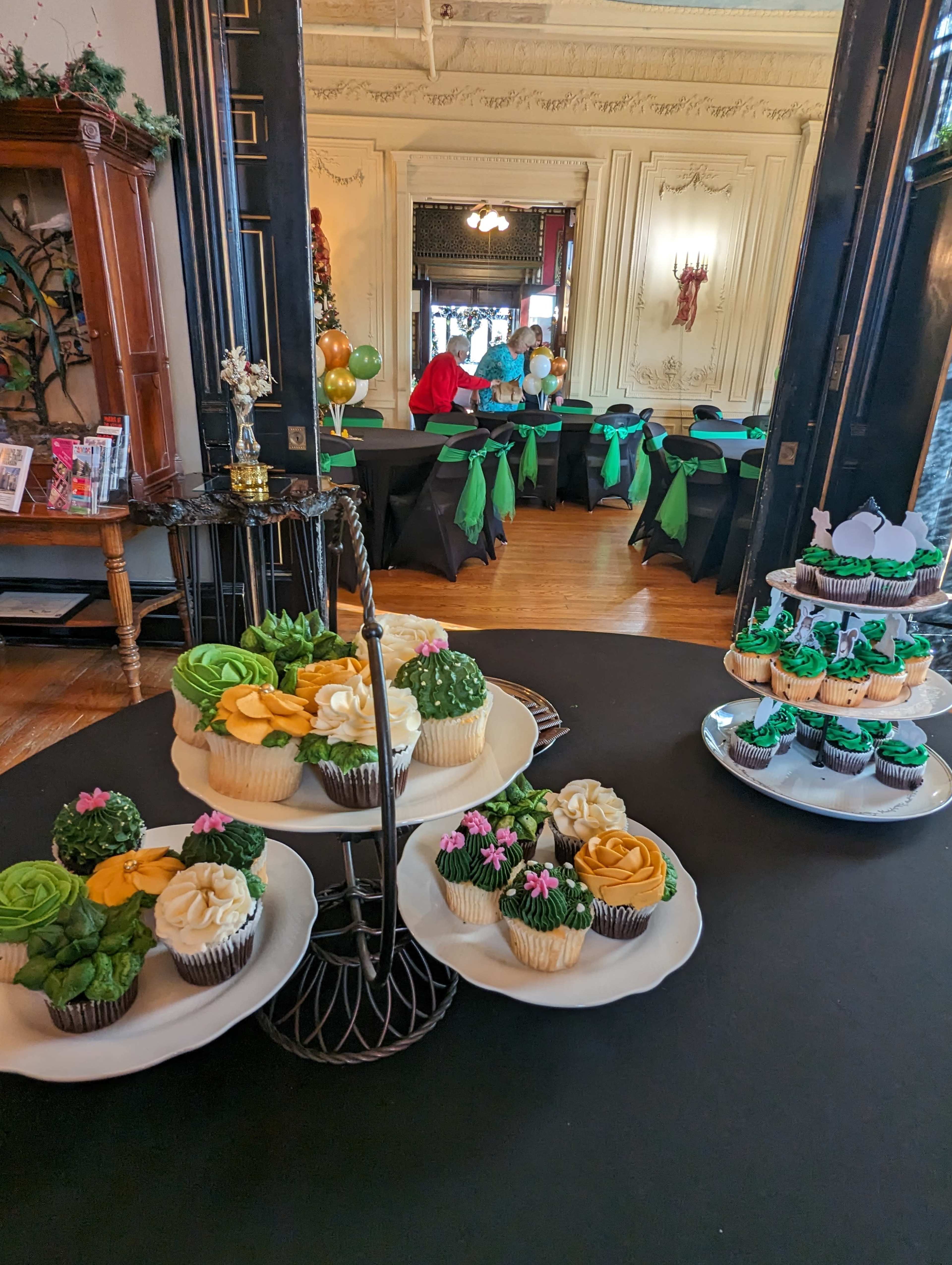 The image features a display of decorative cupcakes in various colors and designs set on a table at a venue with guests seated in the background.