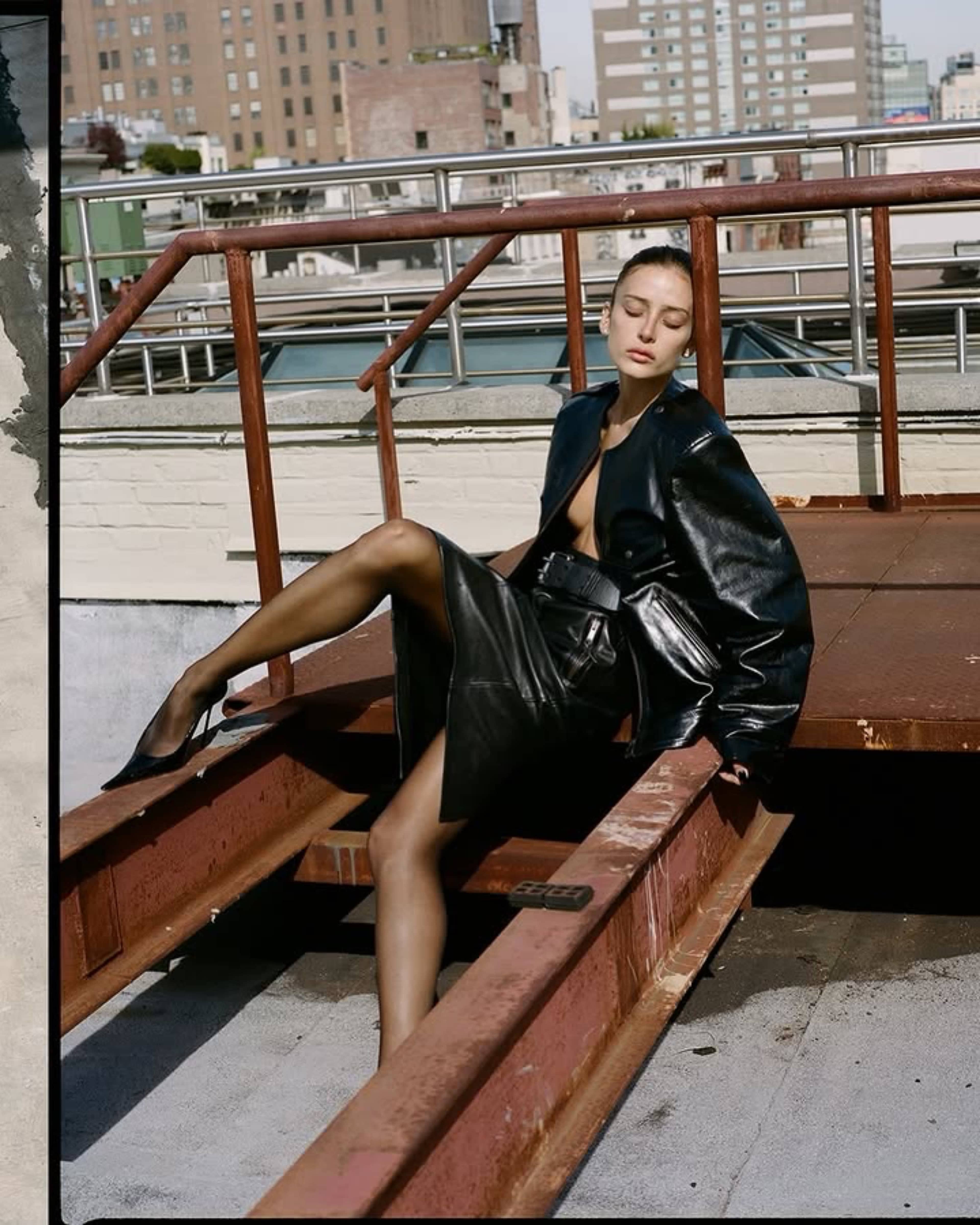 A woman in a black leather outfit sits on a metal framework on a rooftop, with city buildings in the background.