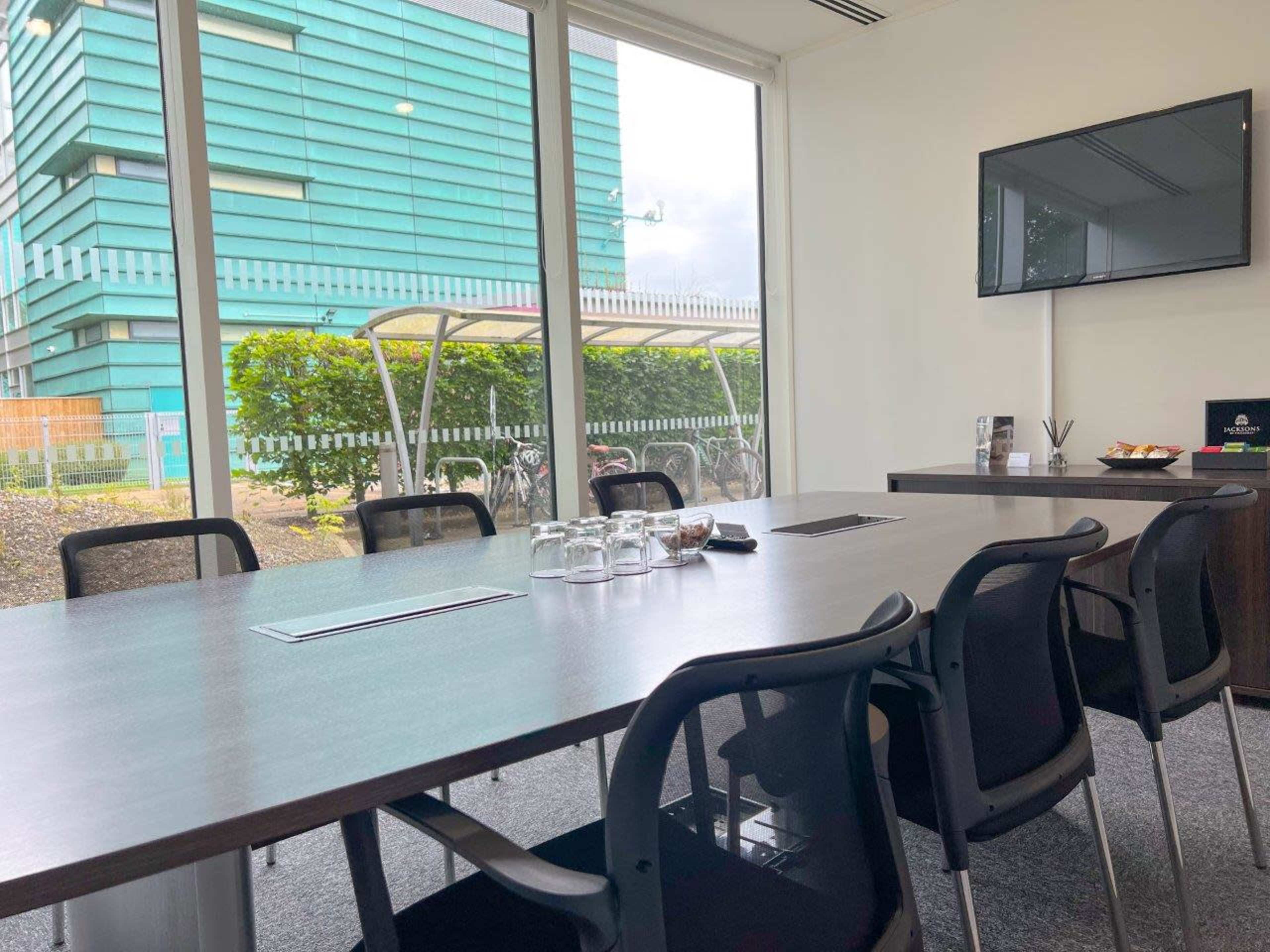 A modern conference room features a large wooden table surrounded by chairs, with large windows overlooking an outdoor area.