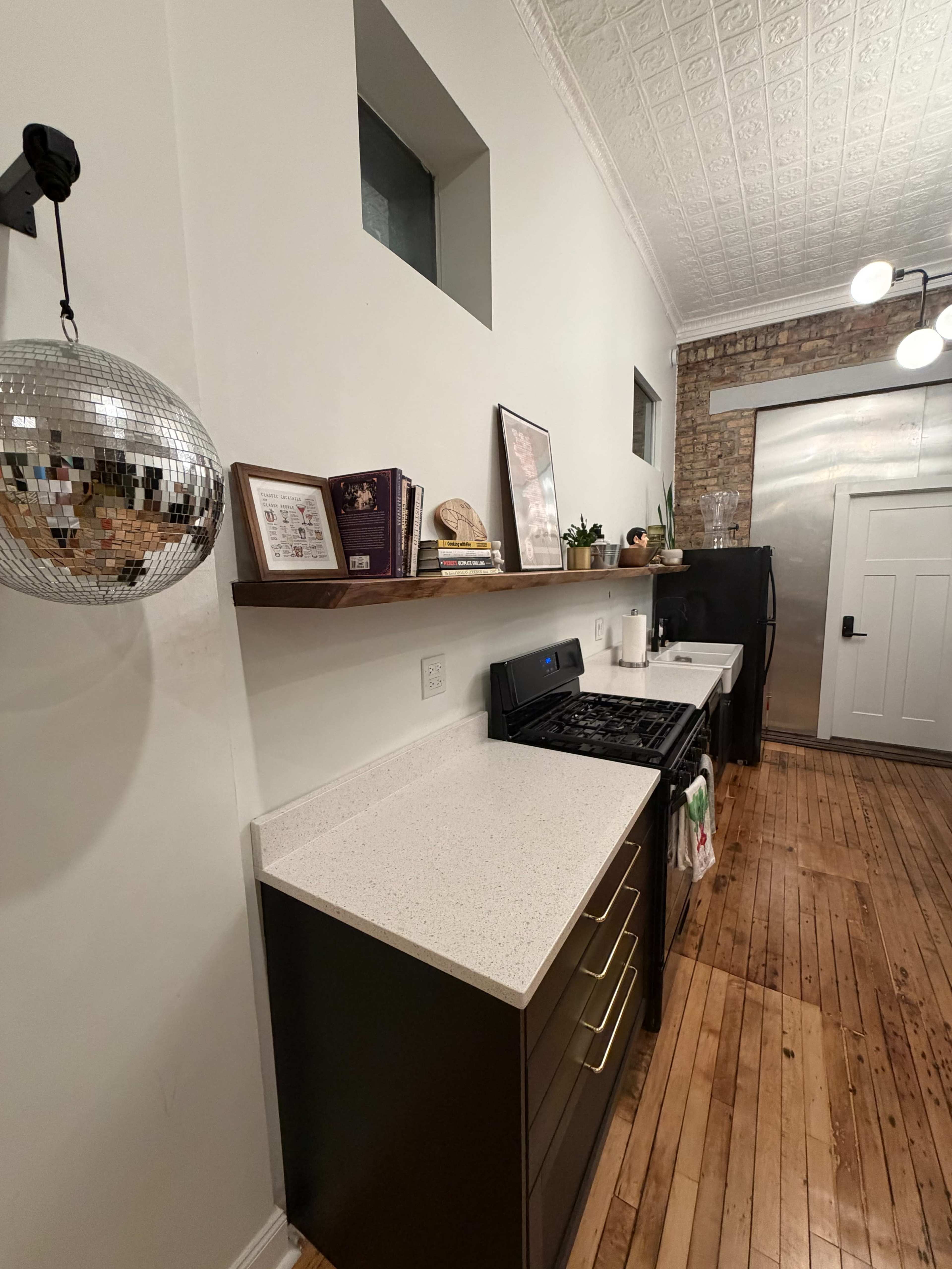 A modern kitchen features a gas stove, open shelving with cookbooks, and a disco ball suspended on the wall, with wood flooring and exposed brick in the background.