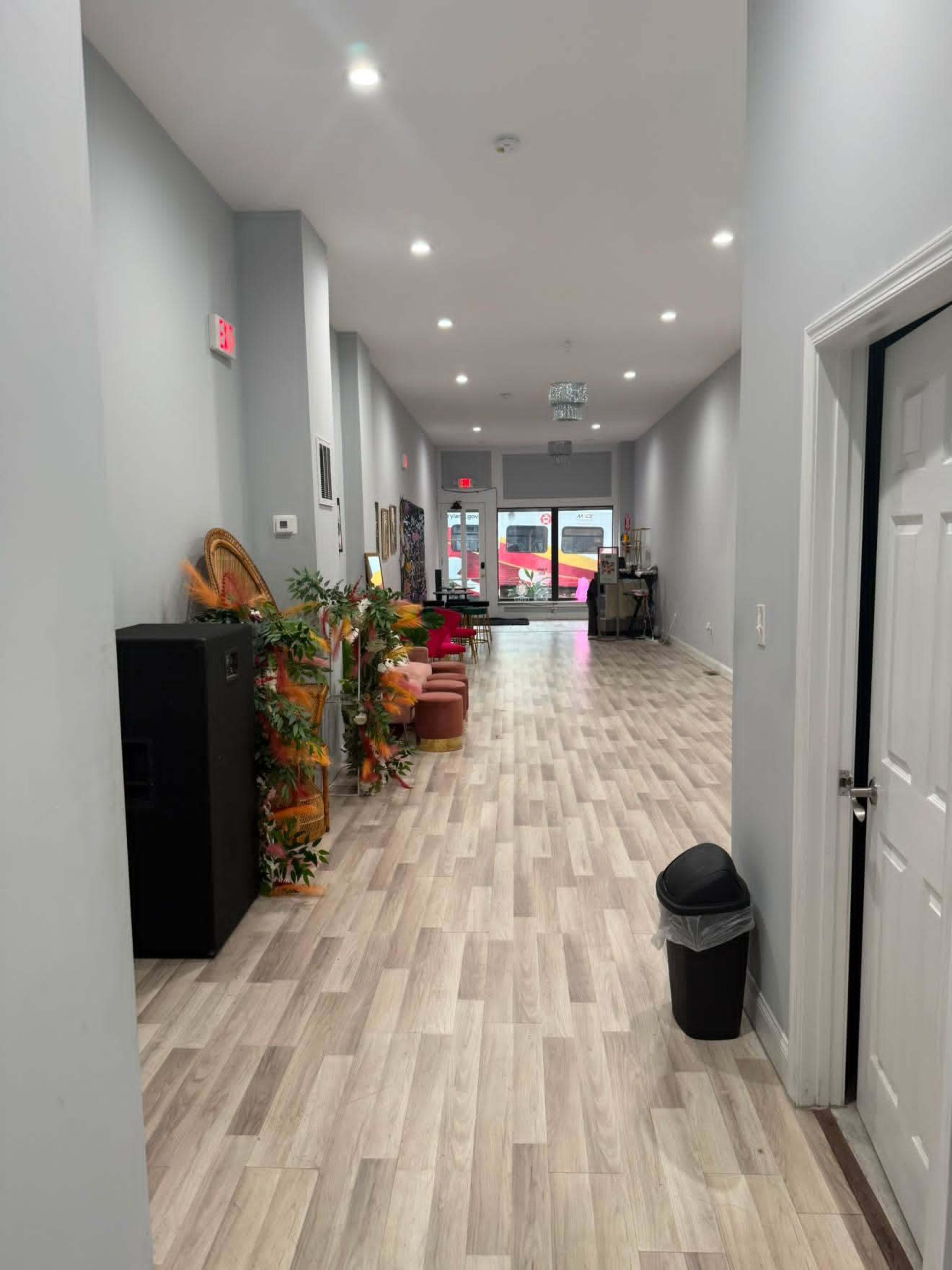A long hallway with gray walls, light wood flooring, and various seating options leads to a bright entrance at the far end.