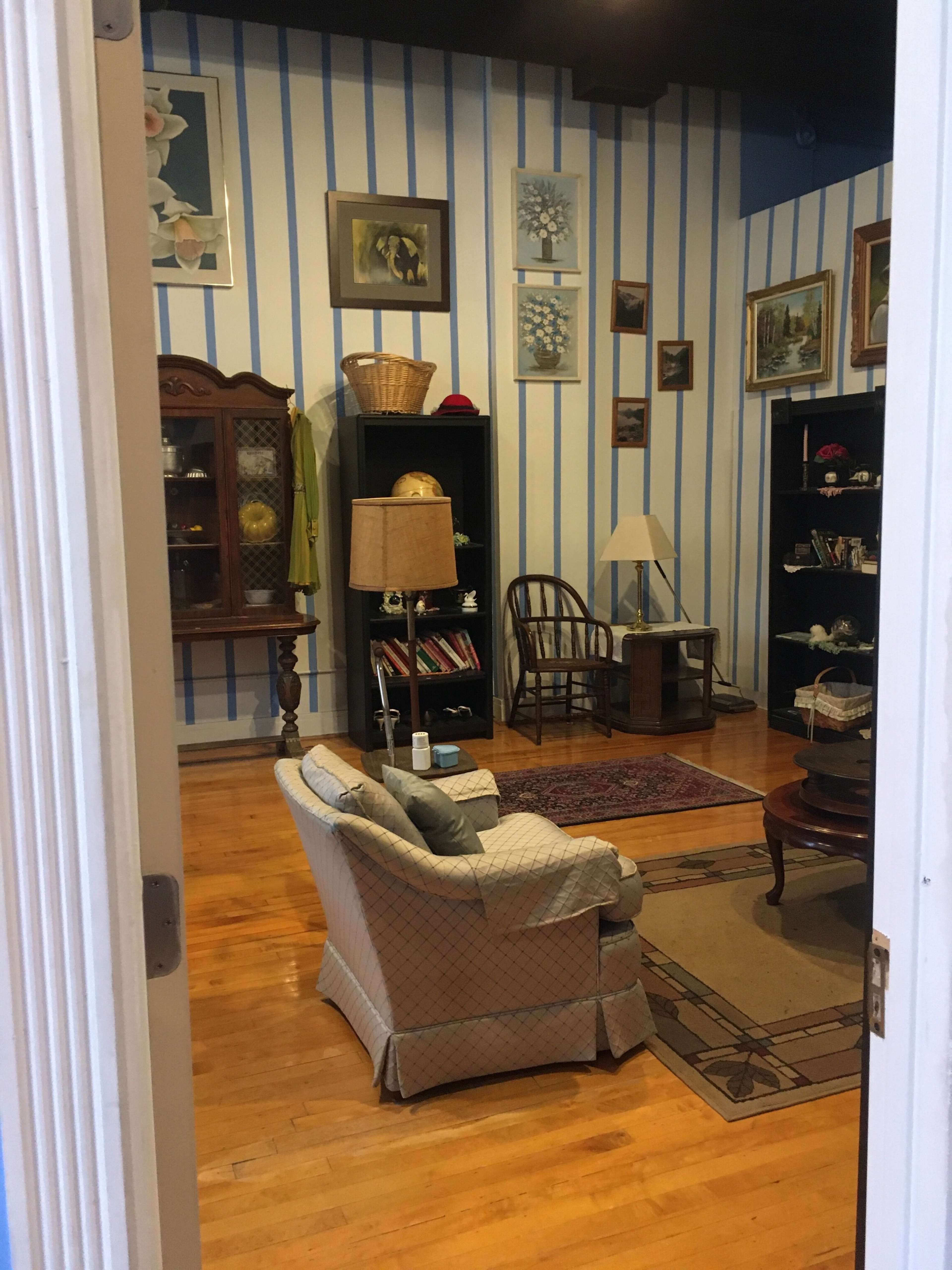A cozy room with blue striped walls, wooden flooring, a patterned rug, and various furniture pieces including an armchair, lamp, and shelves displaying decorative items.