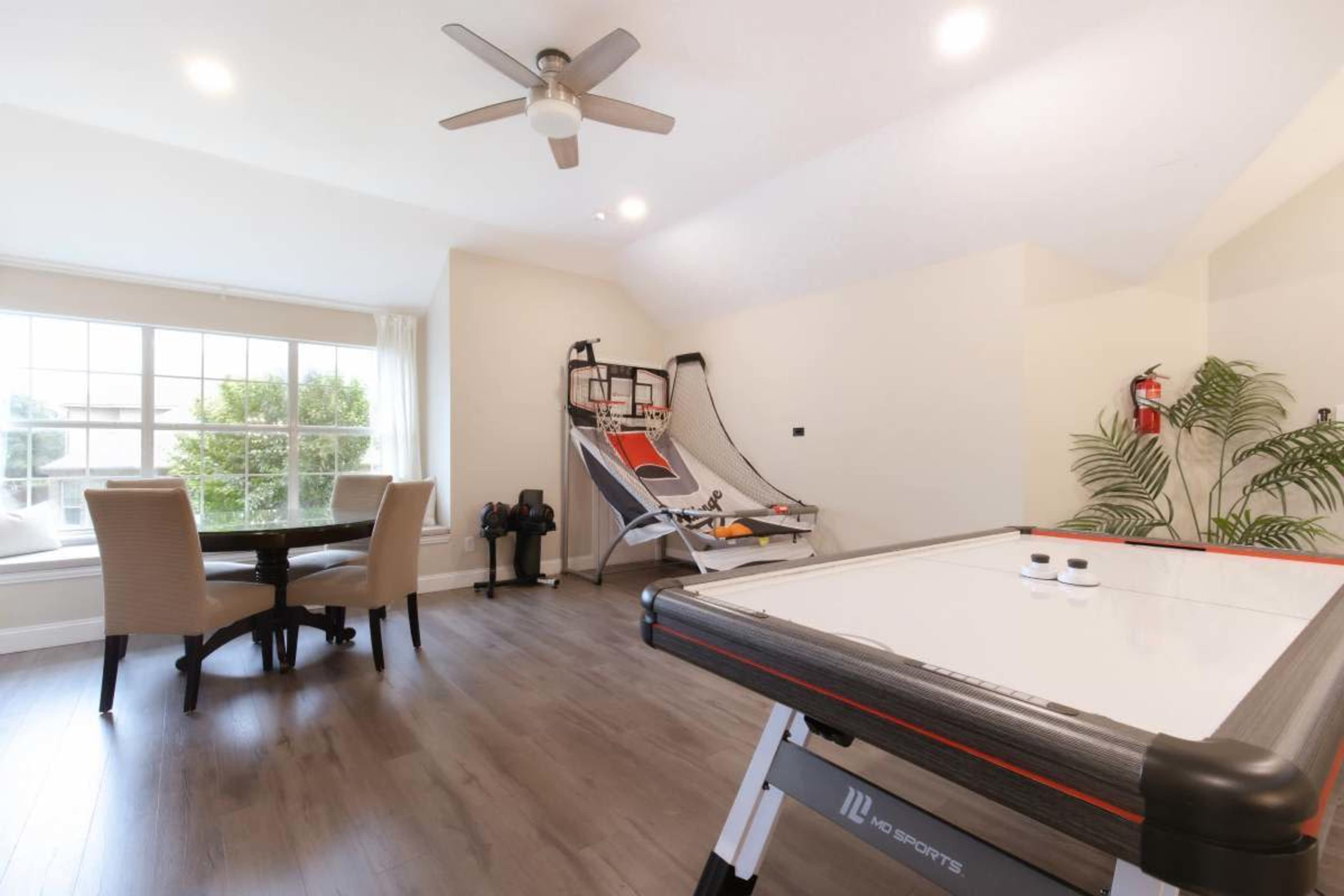 The room features a table for air hockey, a sitting area with two chairs around a small table, and a fitness machine positioned near a large window.