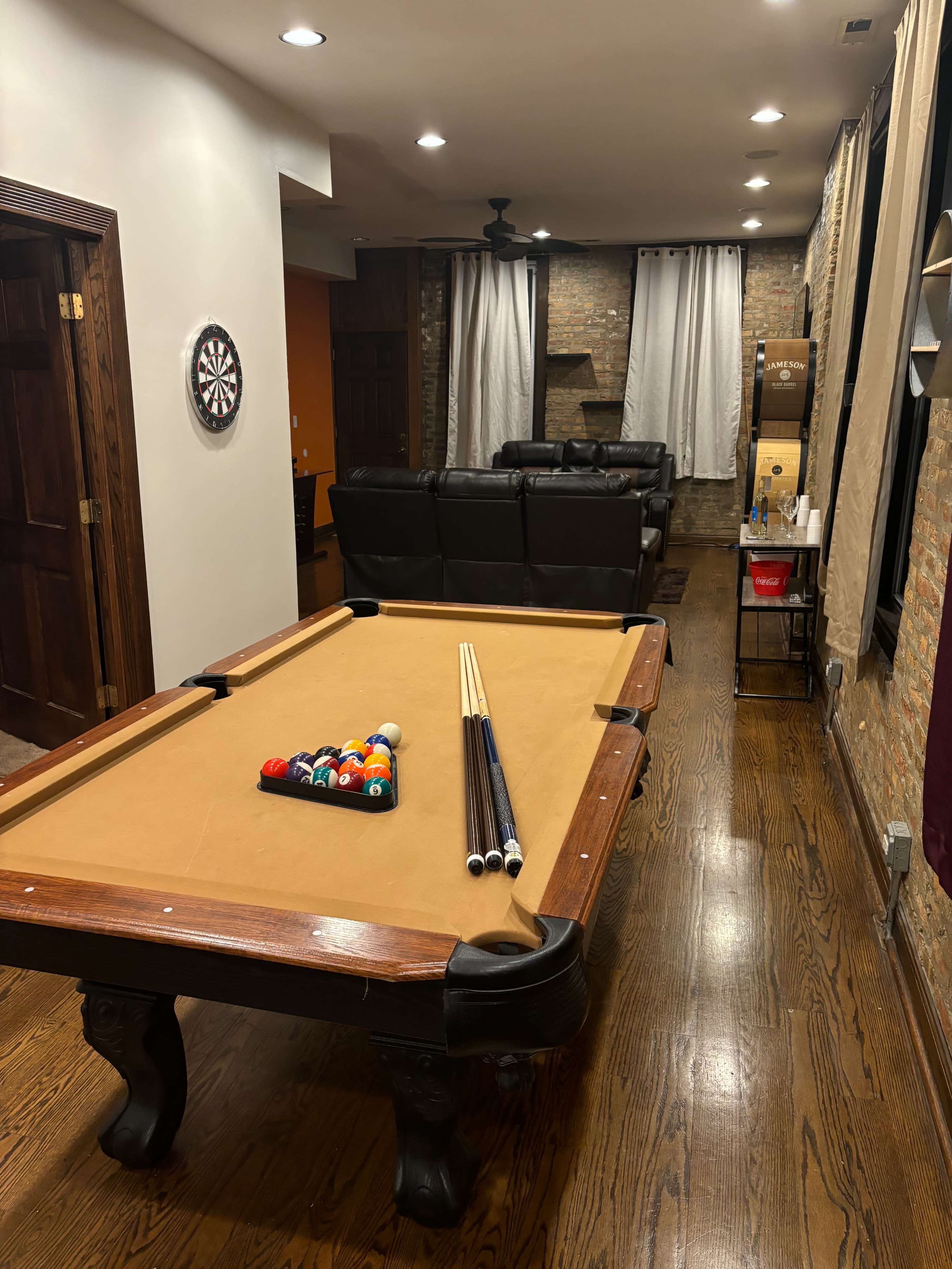 The image shows a billiard table with a triangle of pool balls set up, in a room featuring a comfortable seating area, a dartboard on the wall, and exposed brick accents.