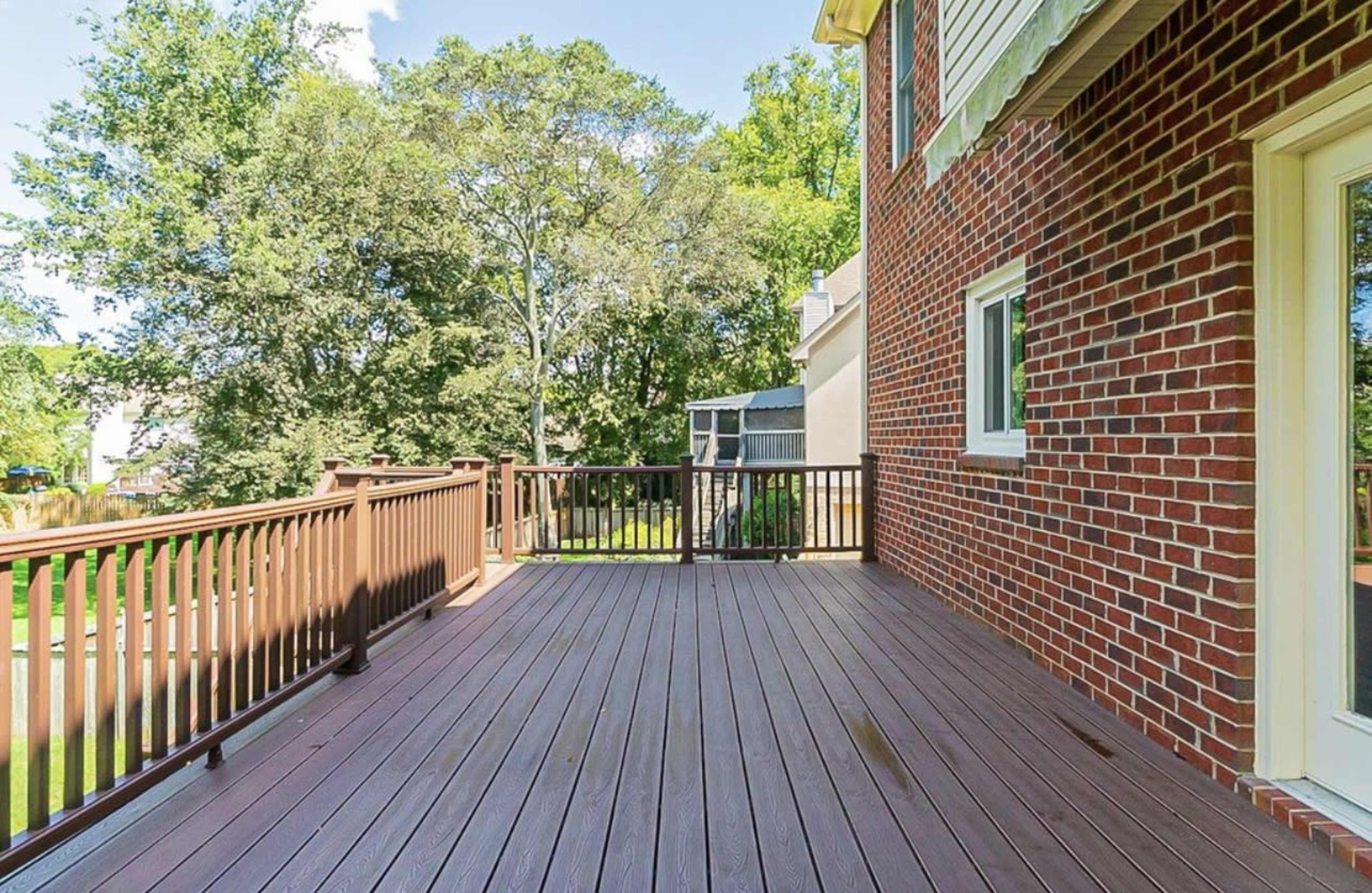 A wooden deck attached to a brick house, surrounded by trees in a sunny backyard.