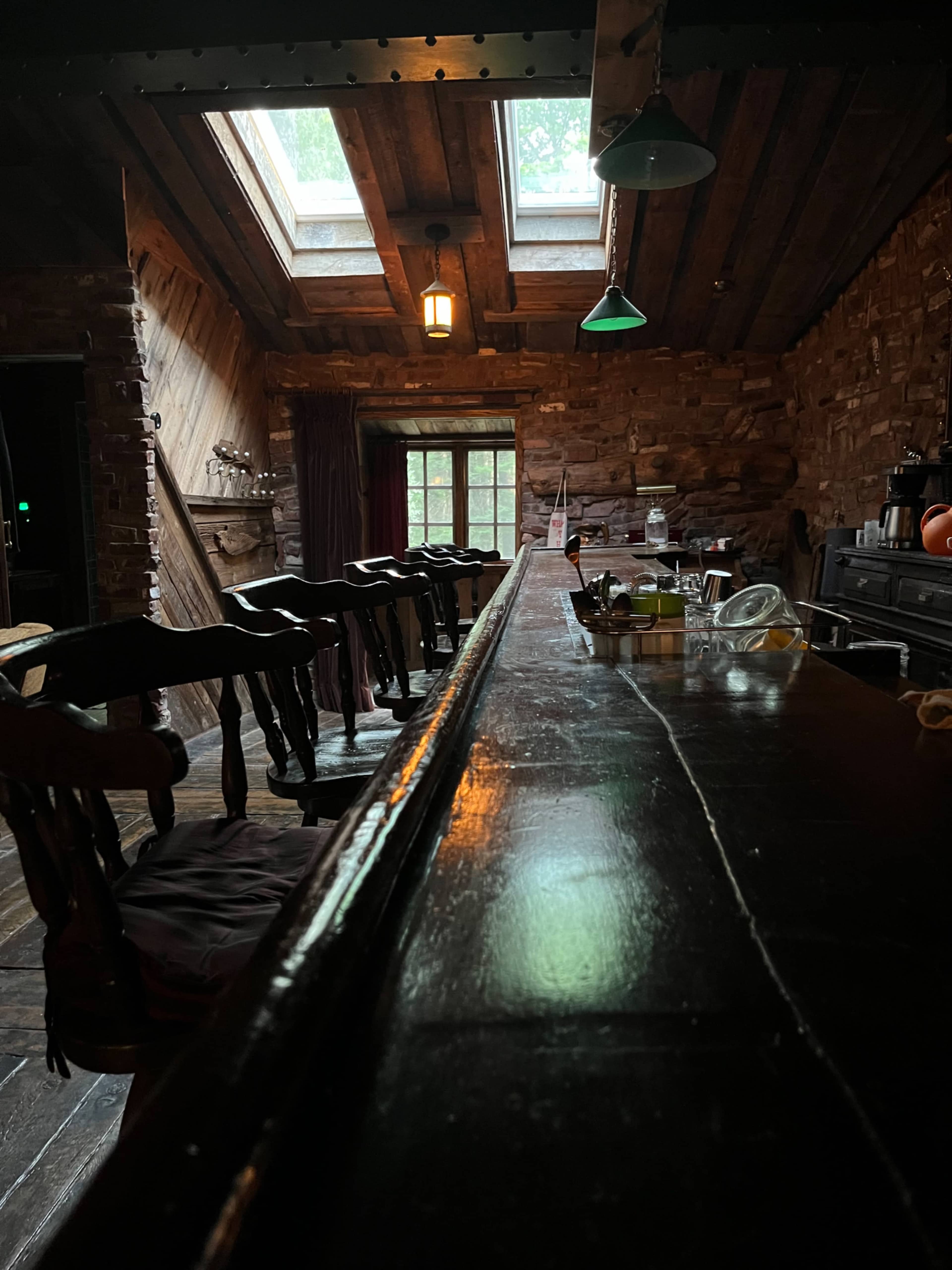 A dimly lit bar with a polished wooden counter, several stools, and a stone wall adorned with various items.