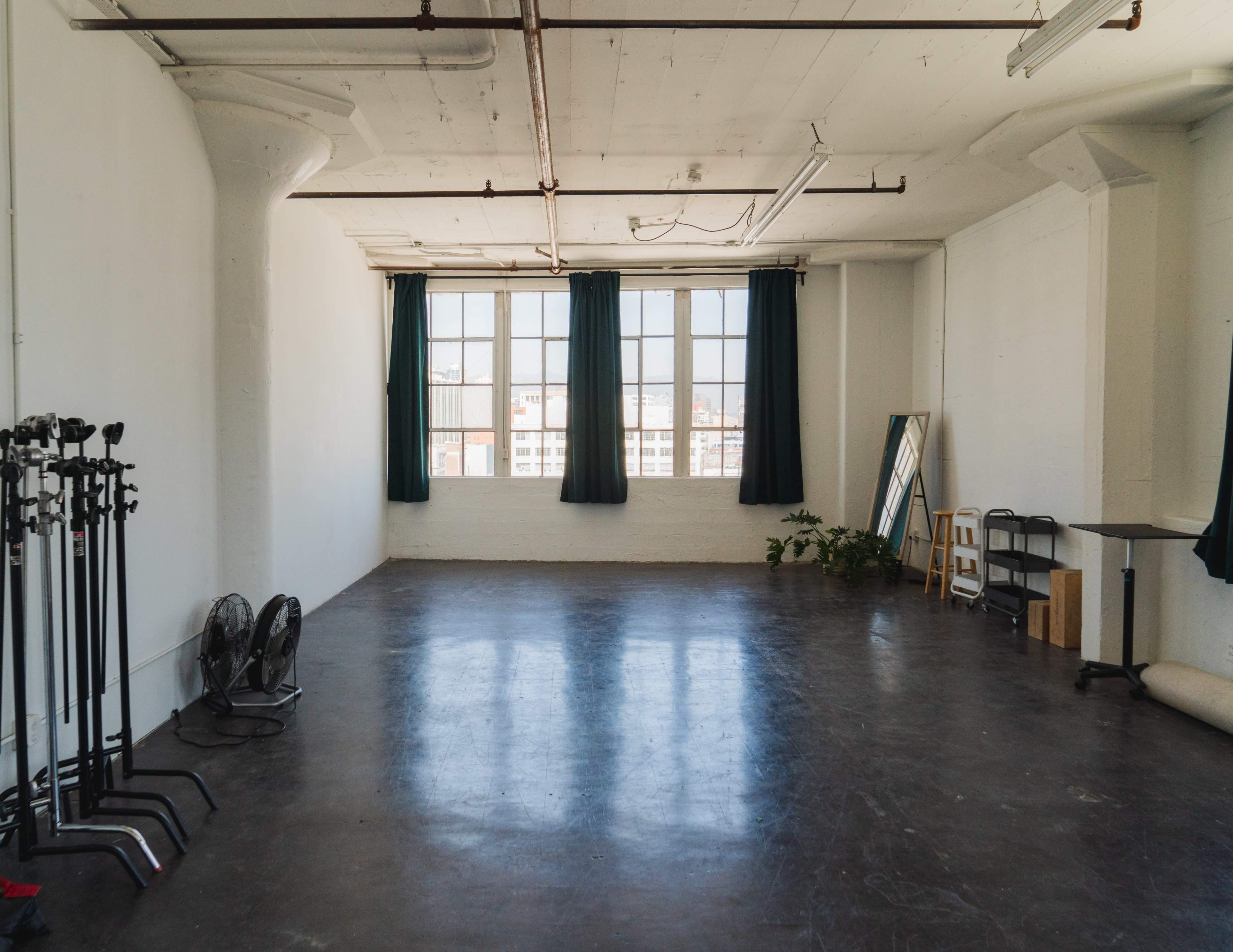 The image shows a spacious, empty room with large windows, green curtains, and several studio equipment stands along one wall.