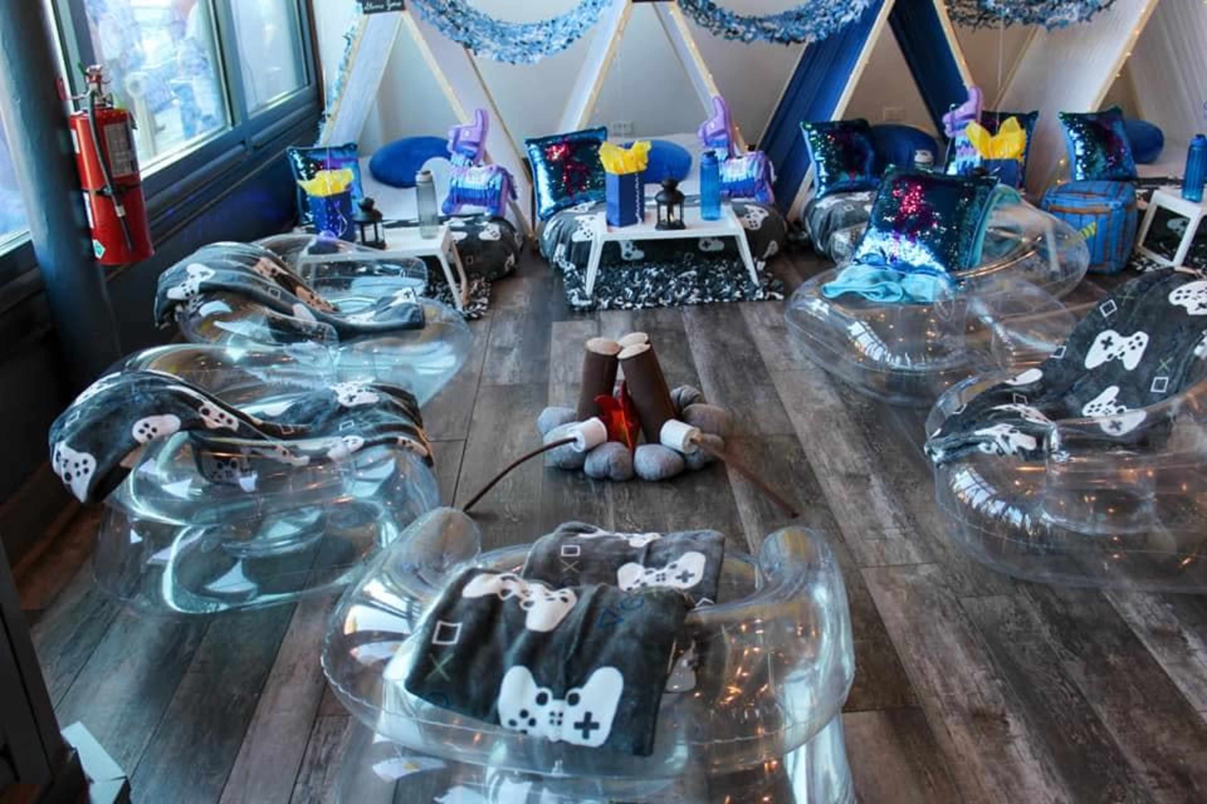 The scene features a modern lounge area with transparent chairs arranged around a circular seating space, decorated with gaming-themed blankets and tables, set in a brightly lit room.