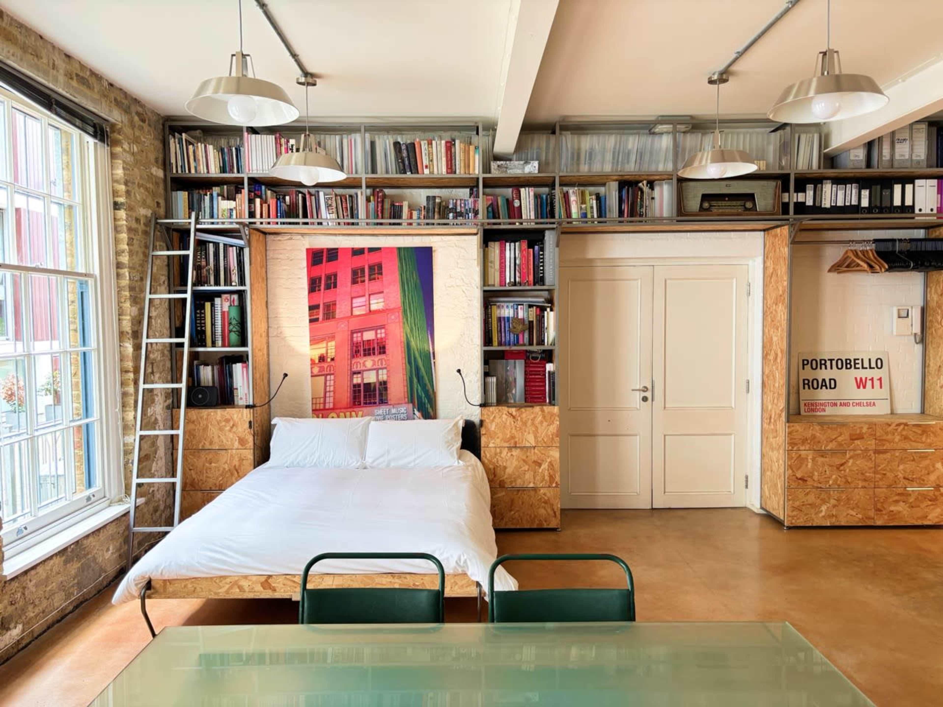 The image depicts a minimalist studio apartment featuring a bed against a backdrop of bookshelves and a large, colorful art piece, with a dining table and chairs in the foreground.