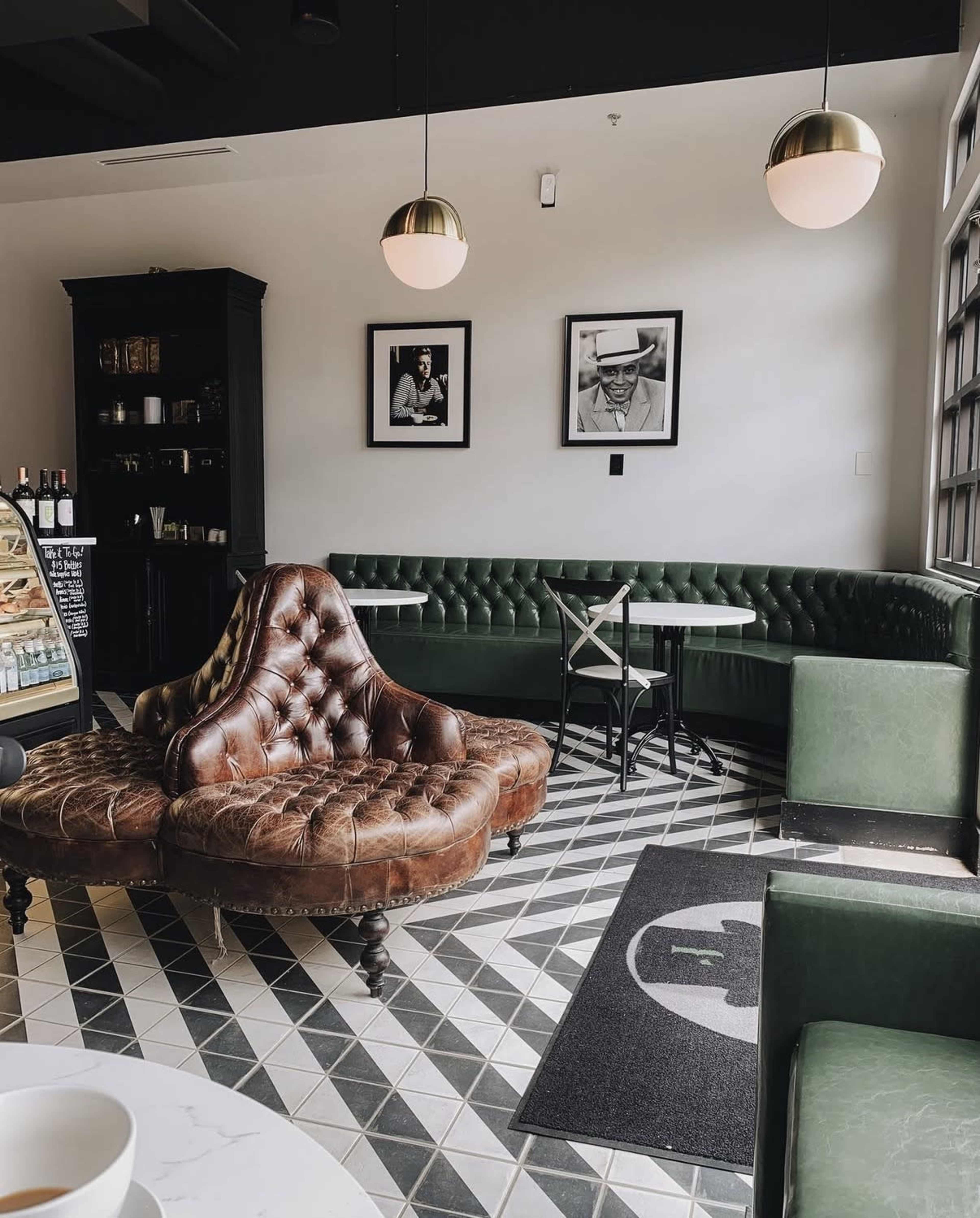 The interior of a café features a brown leather tufted couch, green upholstered seating, and framed black and white photographs on the wall.