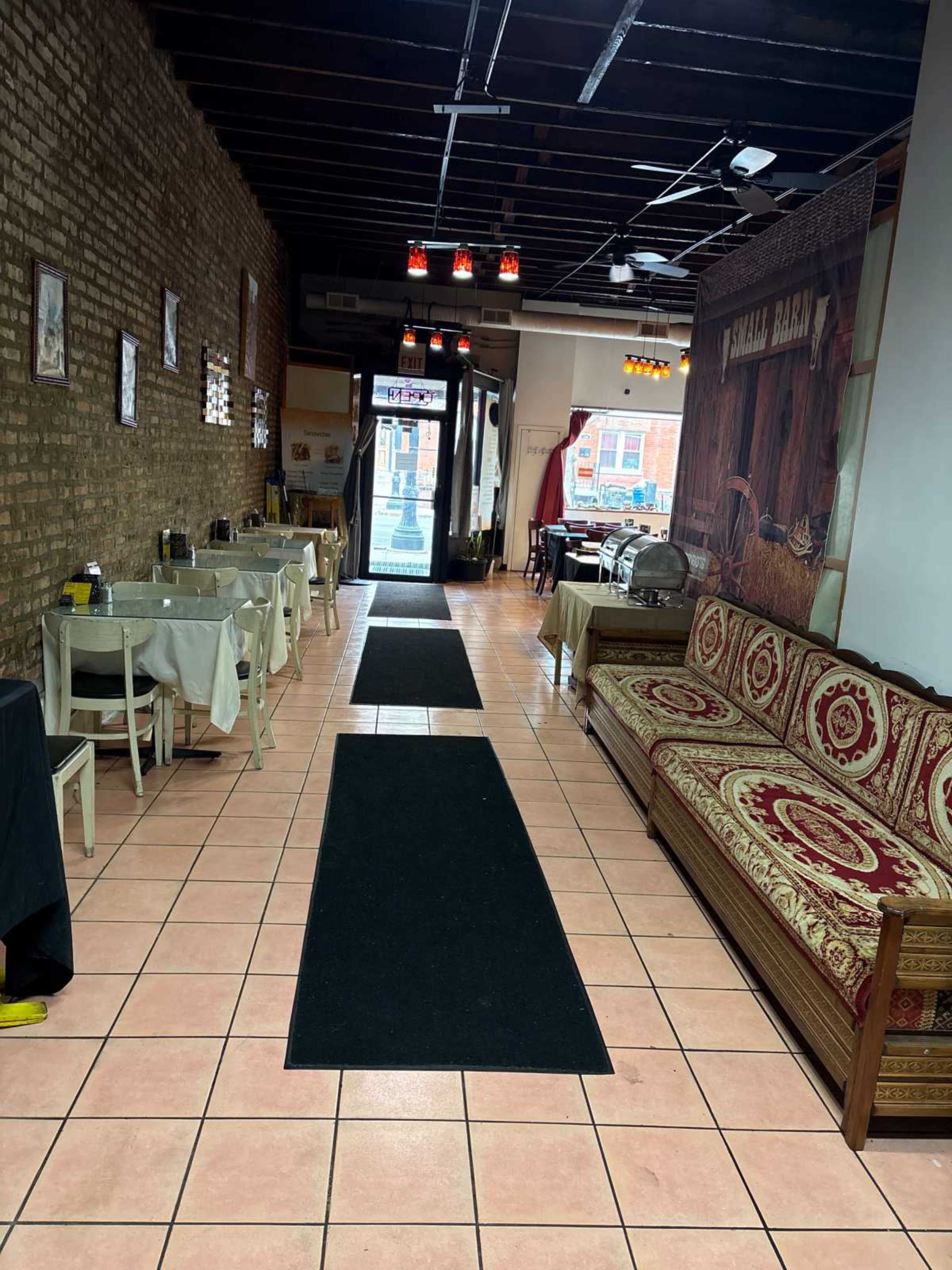 The image shows the interior of a restaurant featuring tiled floors, a long corridor with tables on either side, and a decorative sofa against one wall.
