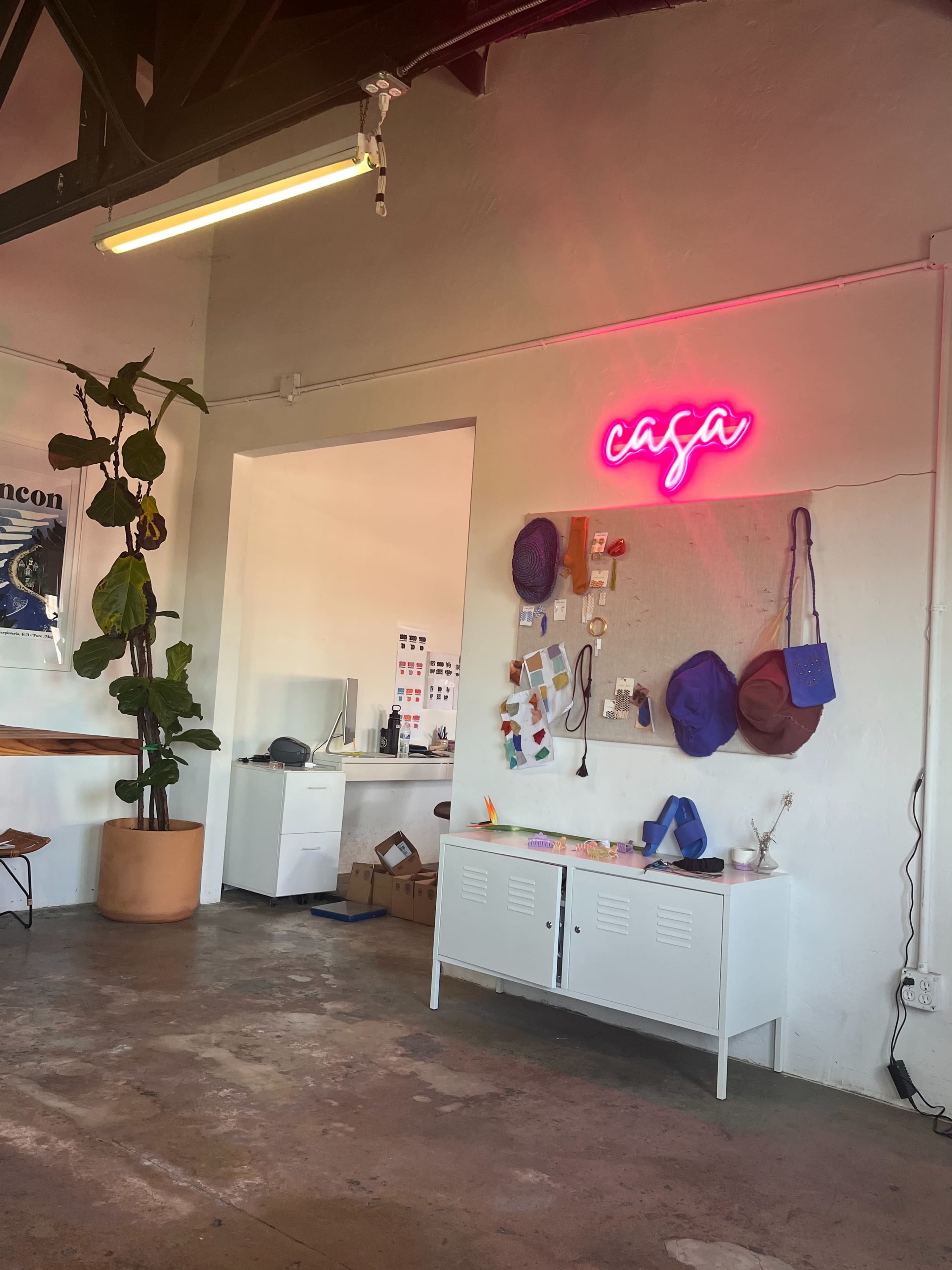 A room featuring a neon sign that says "casa," a plant in a large pot, and a white storage cabinet with various items attached to the wall above it.