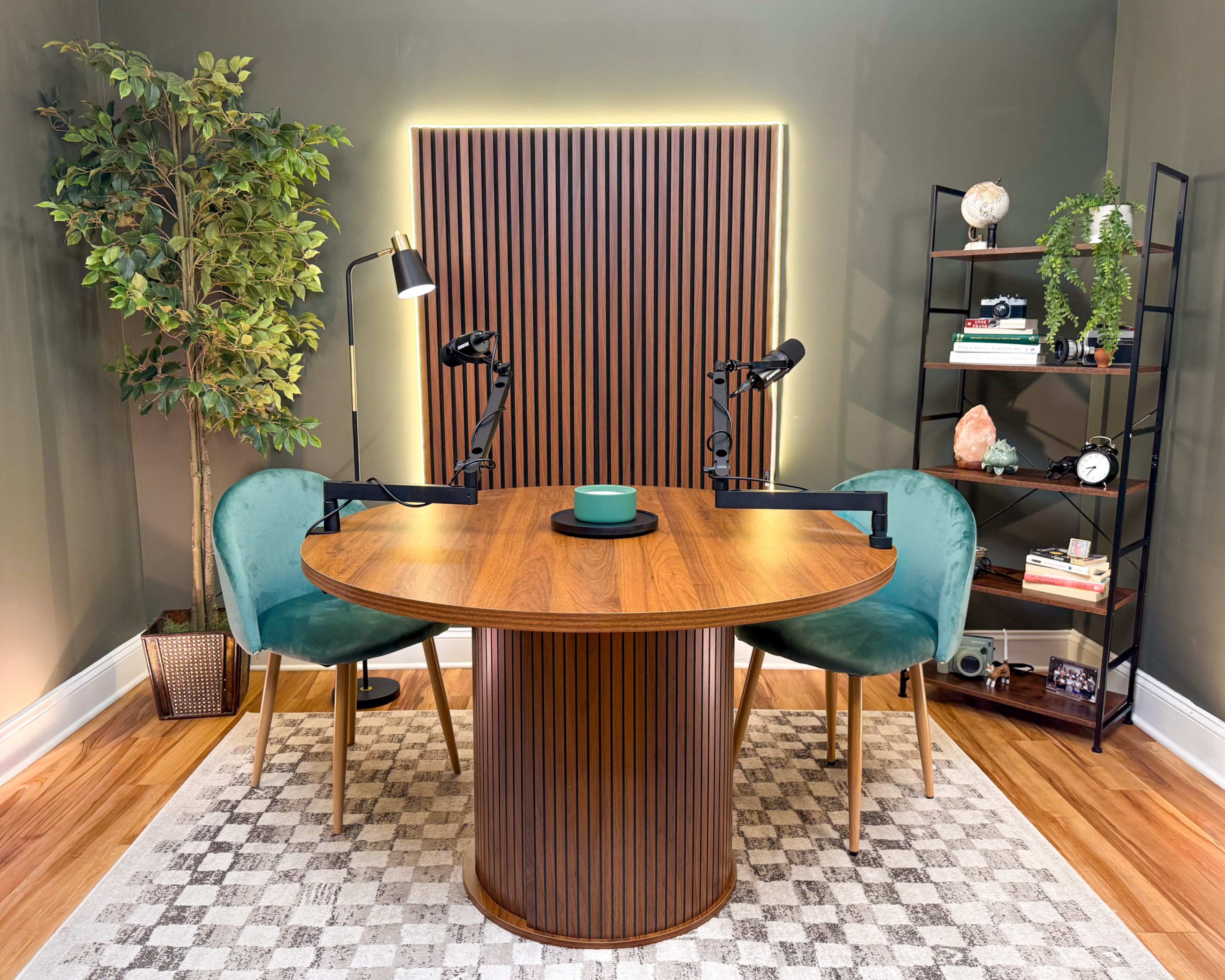 The image shows a modern workspace featuring a round wooden table with two green chairs, surrounded by a plant, a bookshelf, and a wall with vertical wooden slats and ambient lighting.