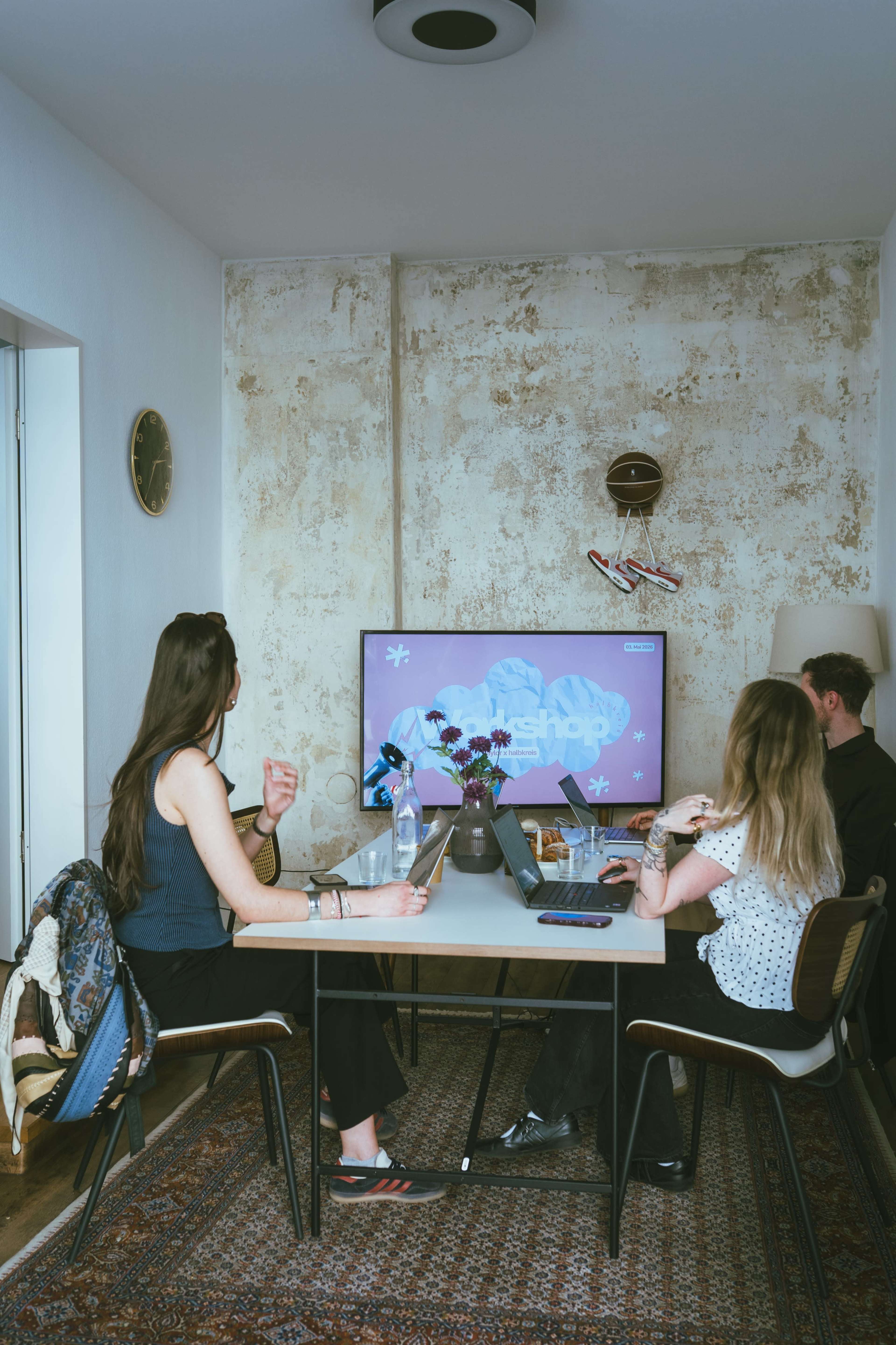 Three people sit around a table in a small room, focused on a presentation displayed on a large screen.