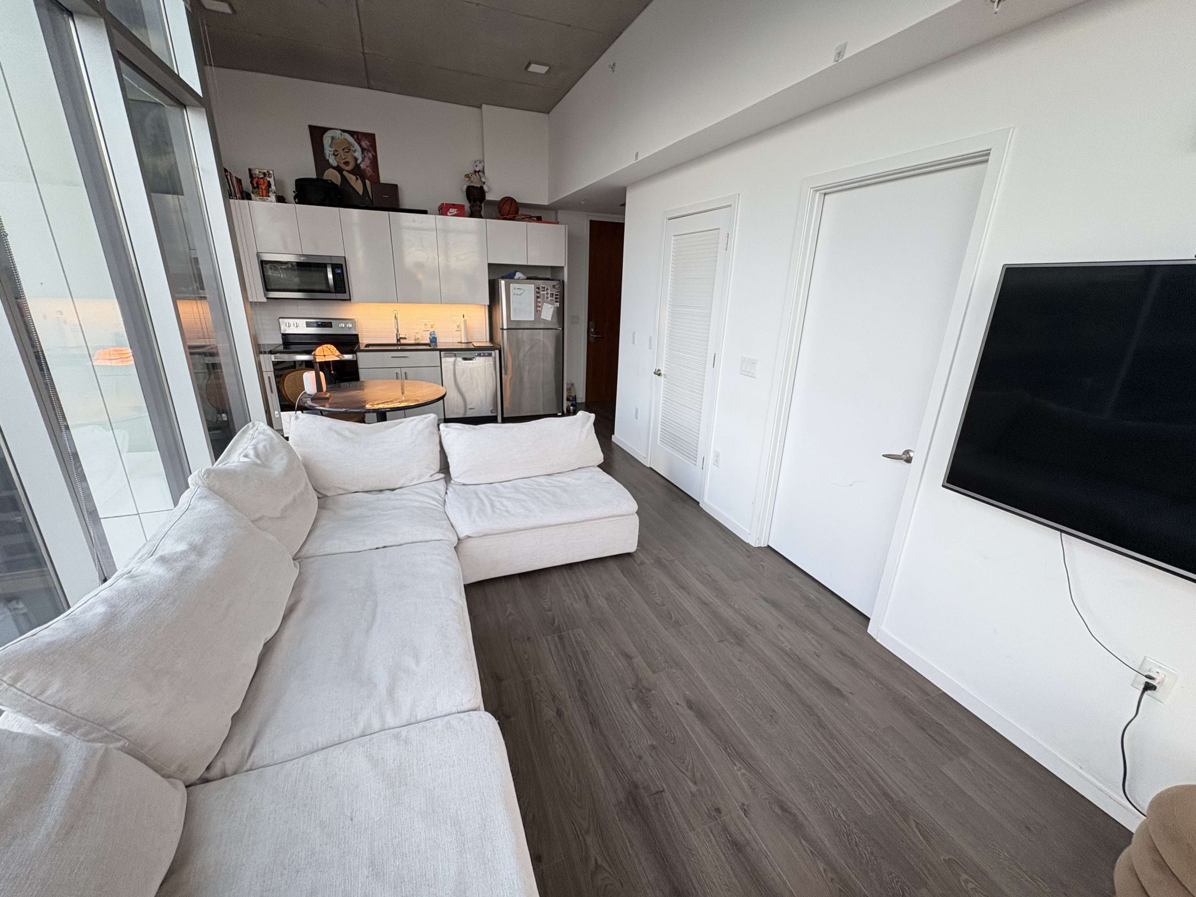 The image shows a modern, open-concept living space with a white sectional sofa, a kitchen area, and a wall-mounted TV.